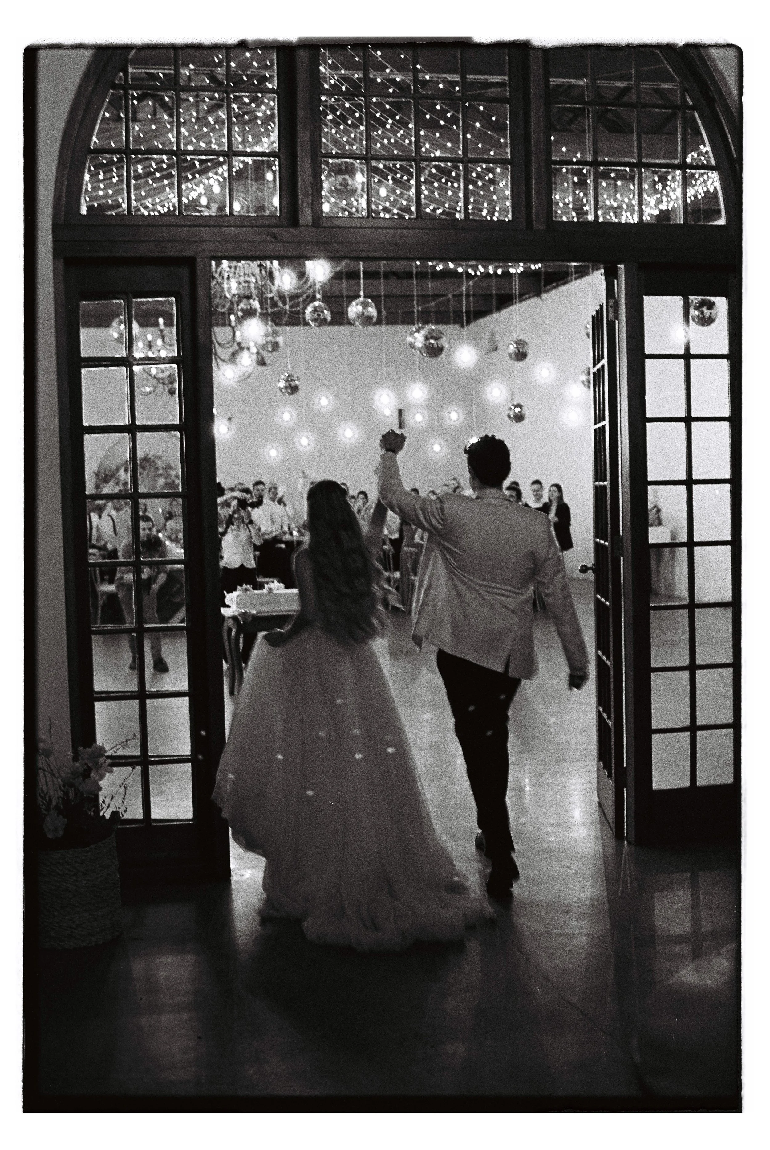 Black and white film photo of bride and groom holding hands entering their reception at Nooitgedacht Estate