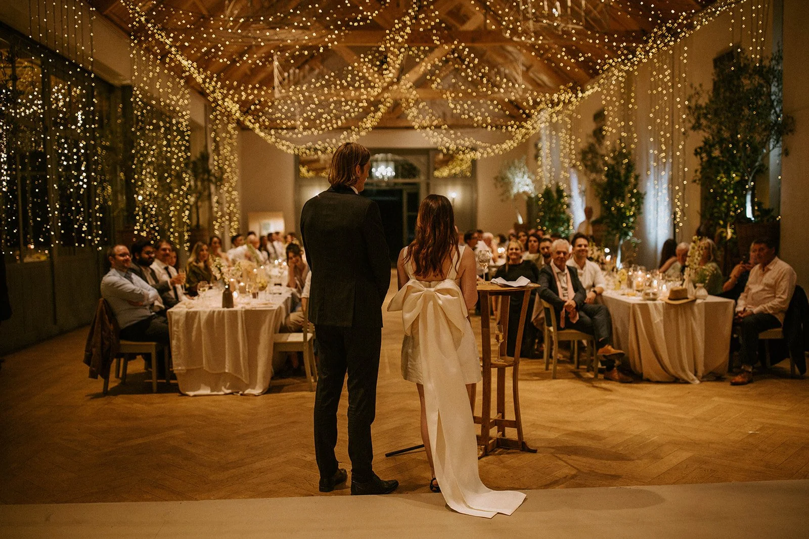 A couple is standing at a wedding reception with guests seated at decorated tables in a beautifully lit, decorated hall with string lights and greenery. The groom is wearing a dark suit, and the bride is in a white wedding dress.