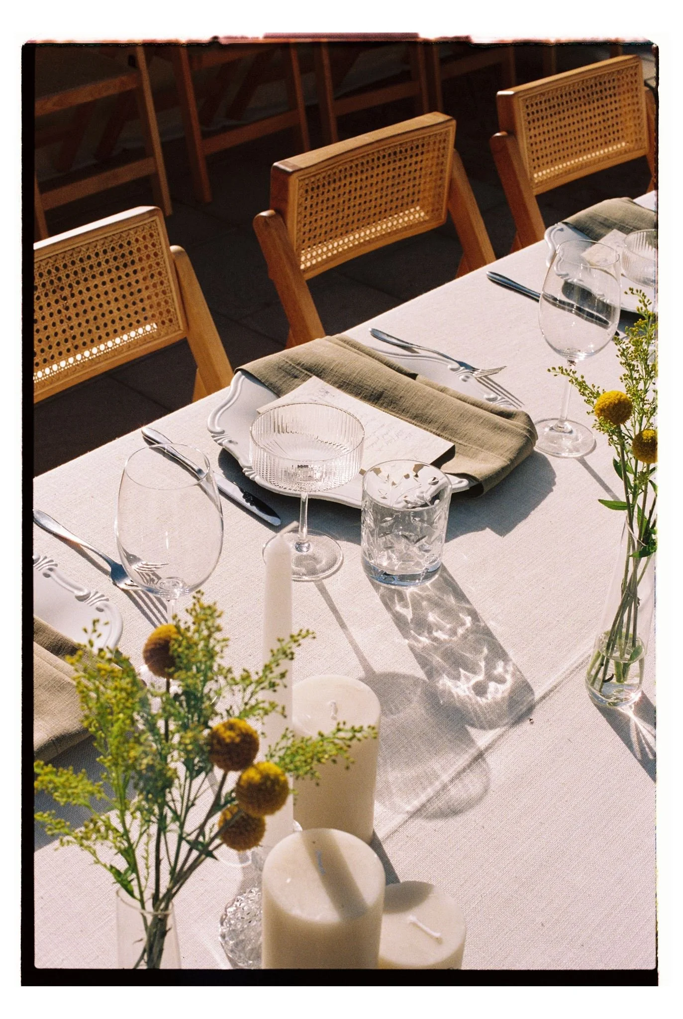 Table set for a formal wedding dinner at Olive Rock with white tablecloth, clear glassware, silverware, a green napkin, and small floral arrangements, with wooden chairs in the background. Captured on 35mm film.