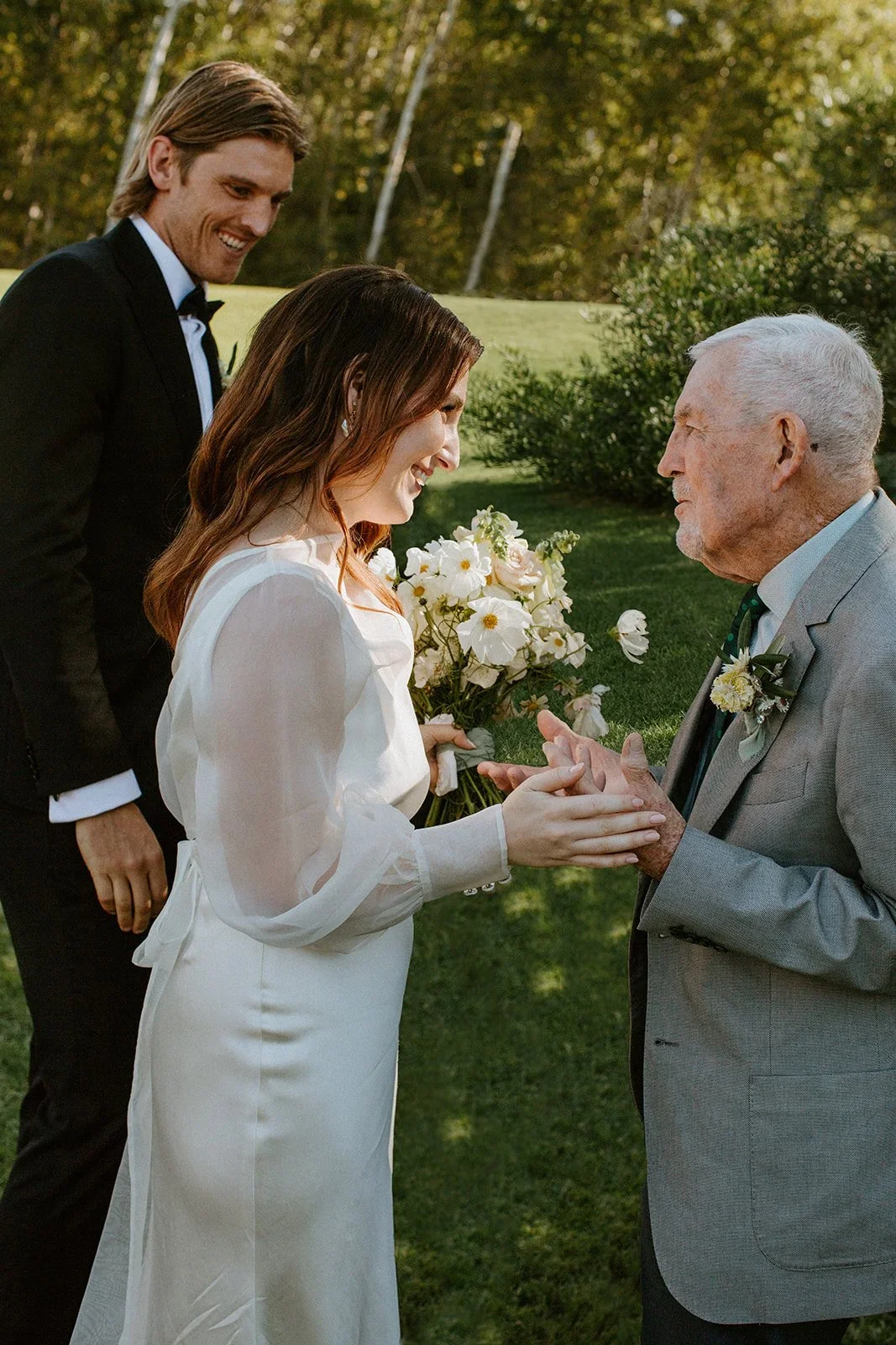 Bride holding her grandfather's hand as she and the groom share a joyful conversation with him after the ceremony at Boschendal Olive Press.