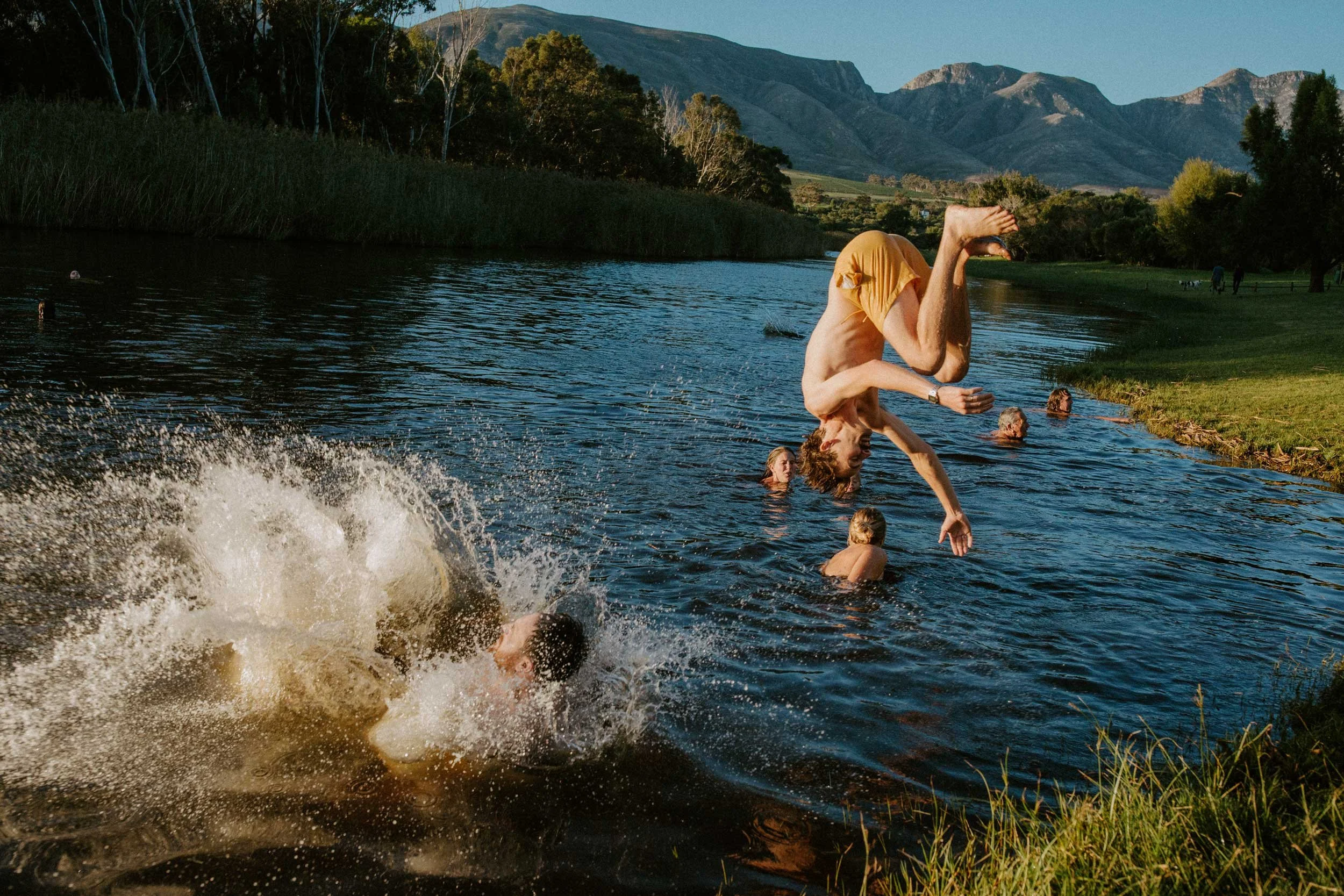 A person in mid-air performing a backflip into a river with other swimmers and a scenic mountain landscape in the background.
