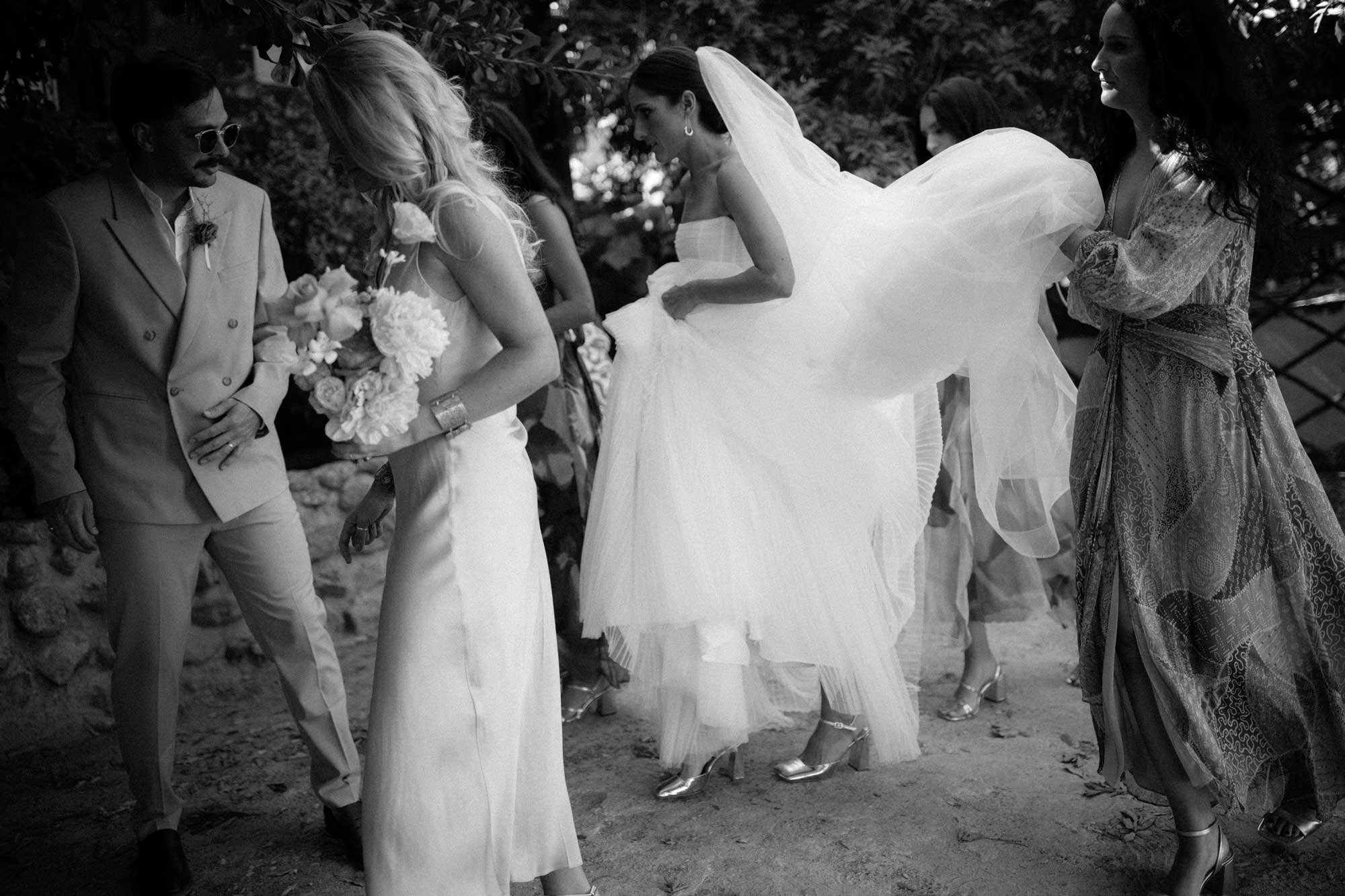 Black and white photo of a wedding scene outdoors with bridesmaids in dresses and heels, a bride in a white gown holding a bouquet of flowers, and smiling. Wedding in Cape Town, South Africa captured by wedding photographer Marli Koen.