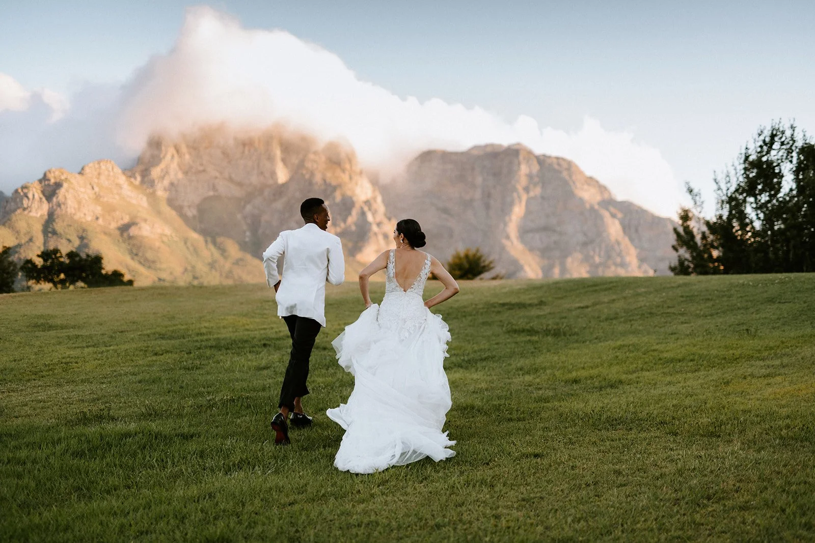A bride and groom running on a grassy field with mountains in the background during sunset.