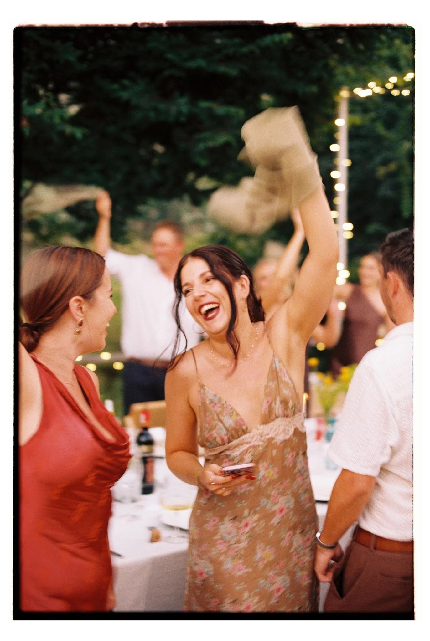 A group of people smiling and chatting at an outdoor social event, with string lights and trees in the background.