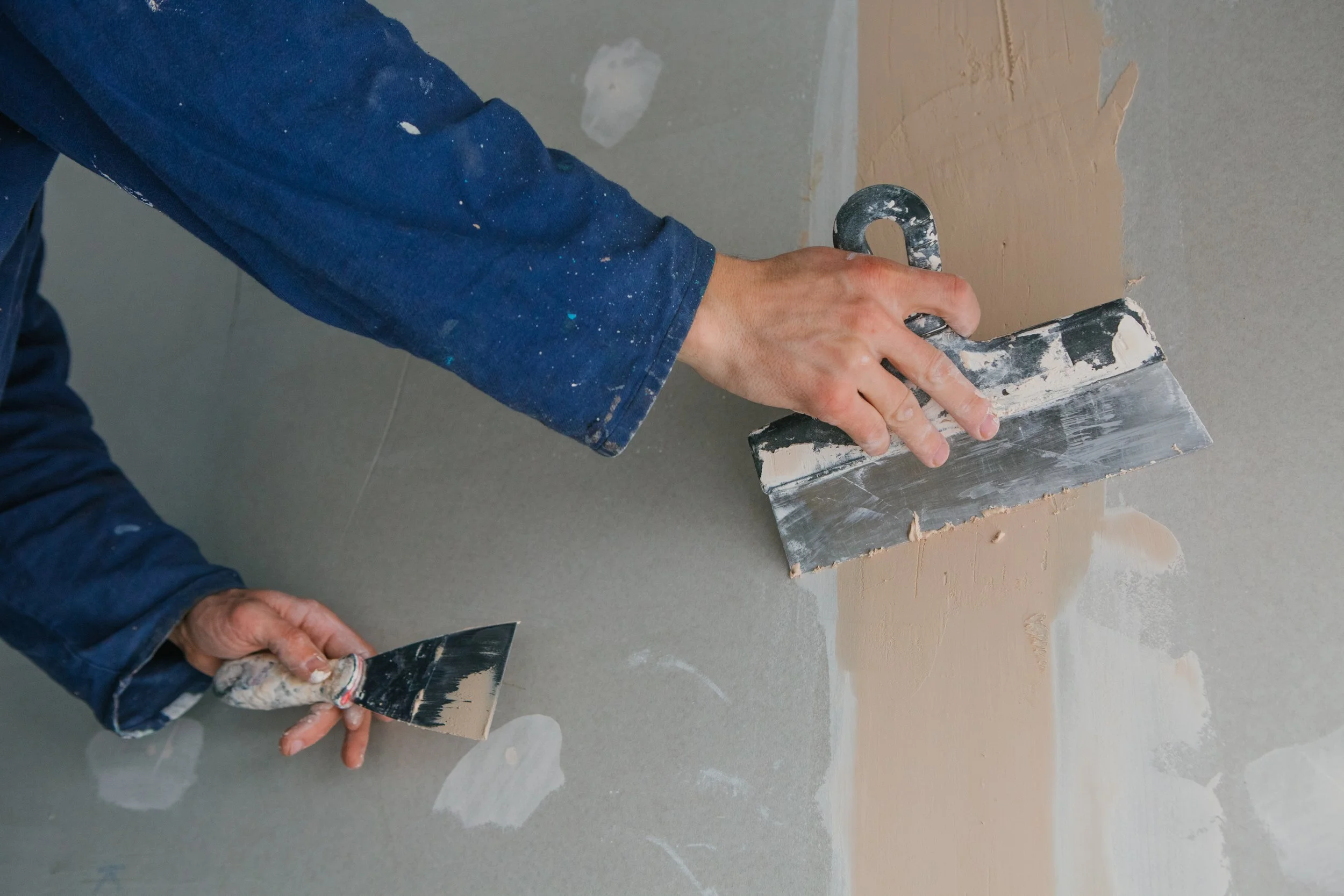 Person wearing a blue sleeve with paint splatters applying a layer of drywall compound to a wall using a putty knife.