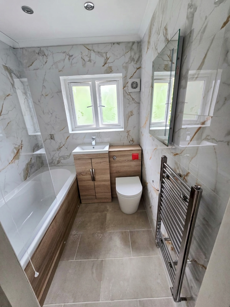 Small modern bathroom with marble walls, a bathtub with a glass partition, a small sink with a wooden cabinet, a toilet, a wall-mounted mirror, a towel radiator, and a window with frosted glass.