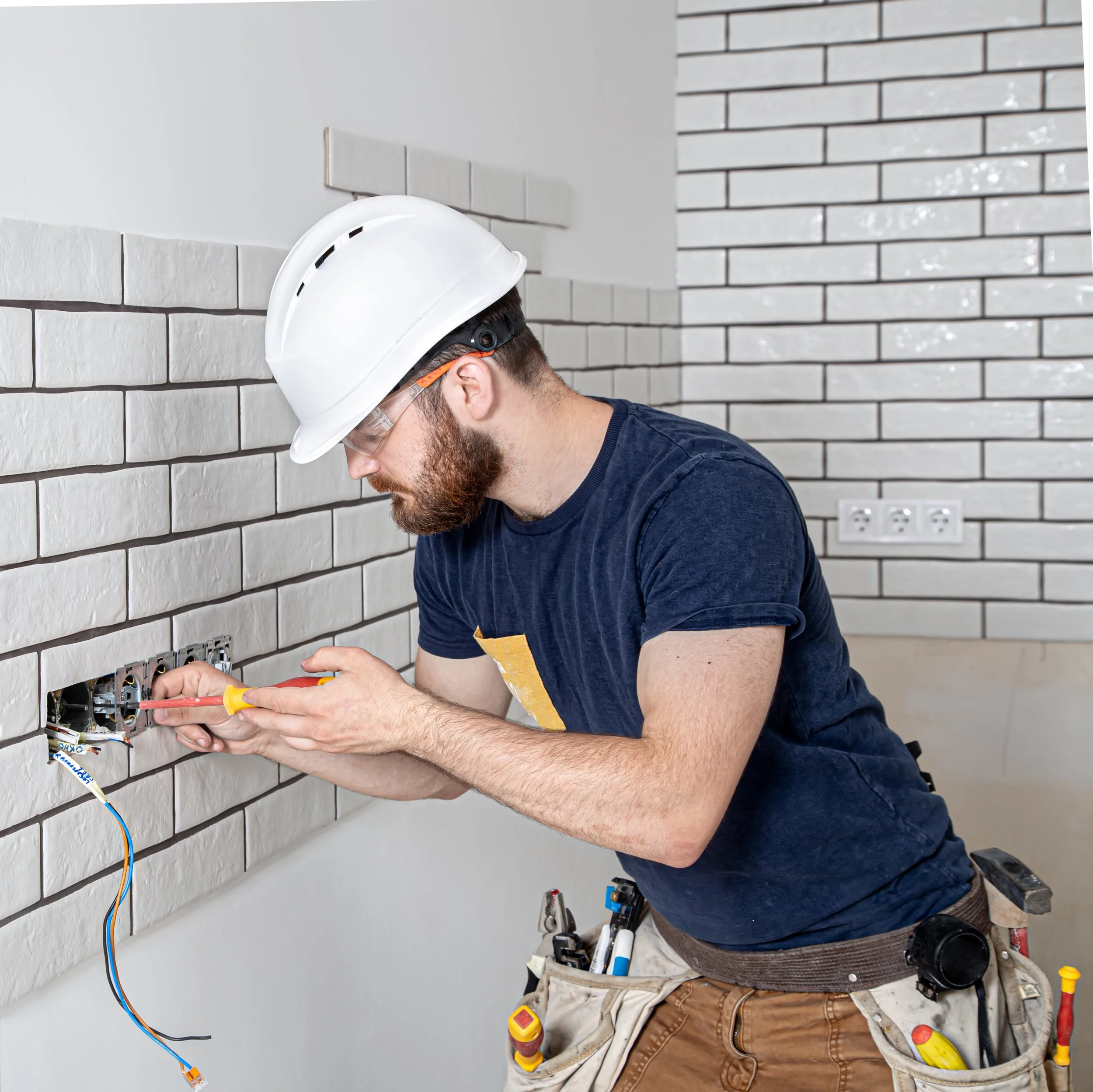 A man working on electrical wiring at a wall, wearing a white safety helmet, glasses, a dark t-shirt, and brown cargo pants with tools hanging from the belt.