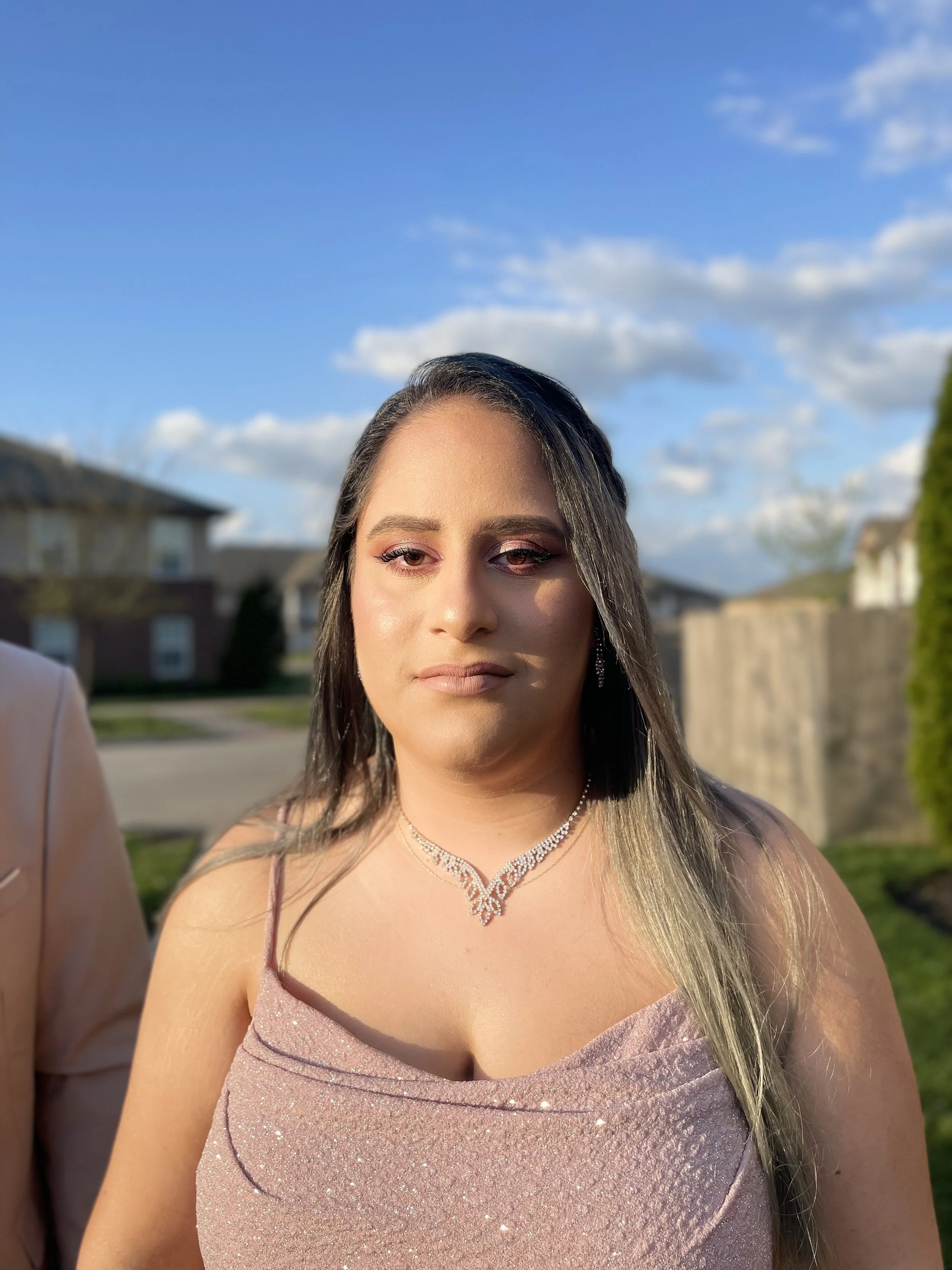 A young woman with long, wavy hair standing outdoors in a pink, glittery dress and diamond necklace, with a background of blue sky, clouds, and houses.