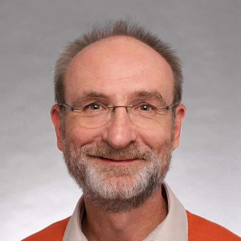 Ein älterer Mann mit Brille, grauem Bart und kurzem Haar, trägt ein orangefarbenes Oberteil vor einem grauen Hintergrund.