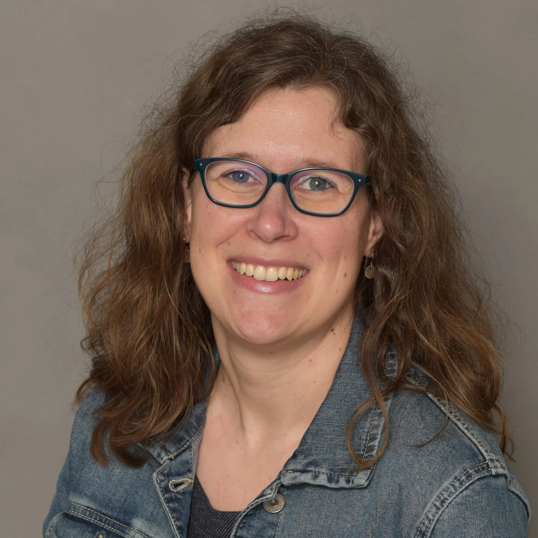 Eine lachende Frau mit hellblauen Augen, braunen lockigen Haaren und Brille, trägt eine Jeansjacke.