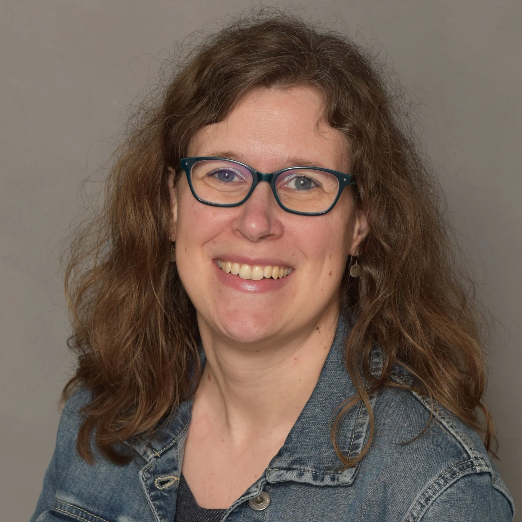 Nahaufnahme einer lachenden Frau mit Brille, braunen, lockigen Haaren, in einem Denim-Jacke vor einem grauen Hintergrund.