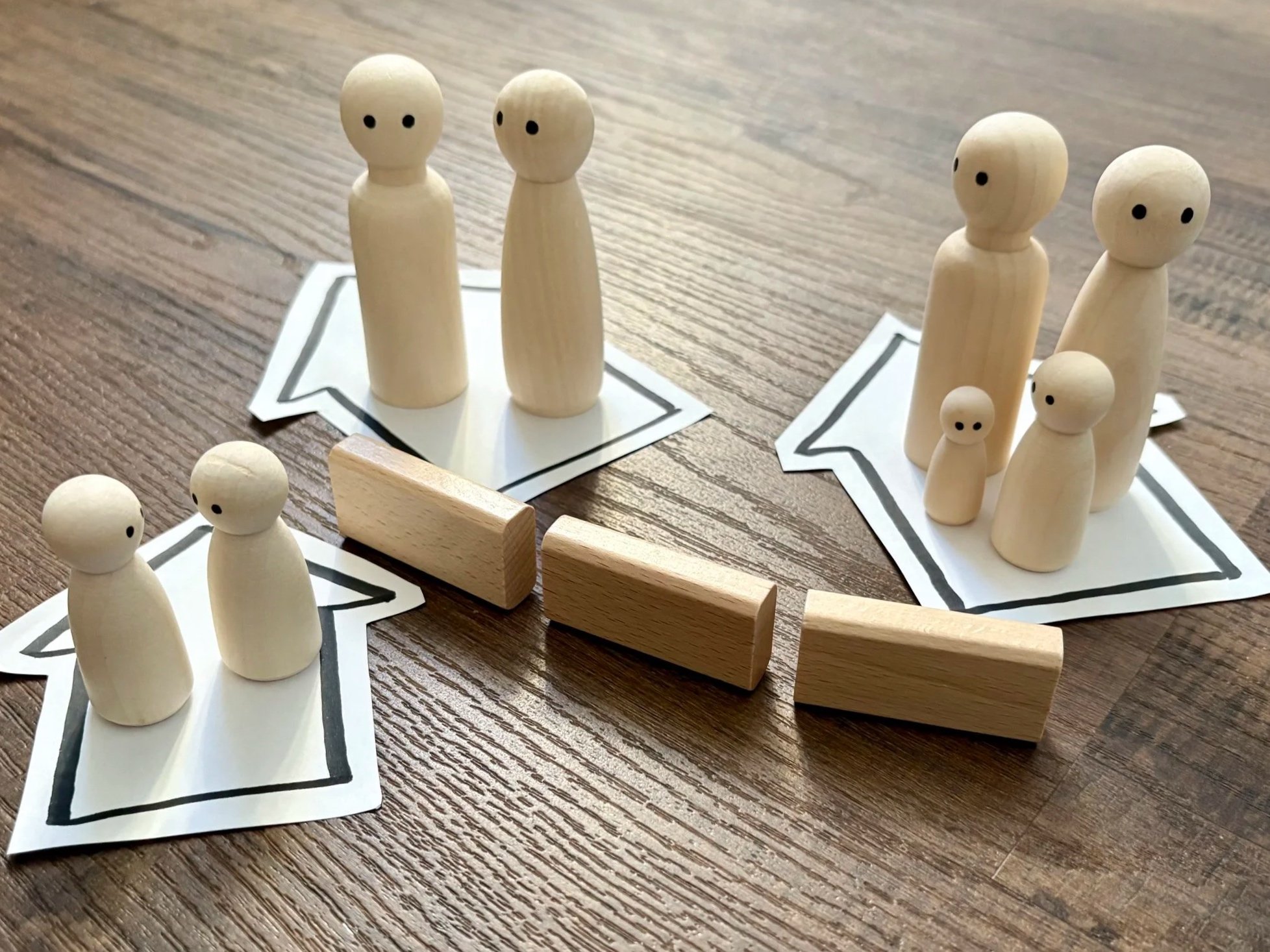 Holzfiguren in Form von Menschen stehen in Gruppen auf Hausformen aus Papier, die auf einem Holztisch liegen.