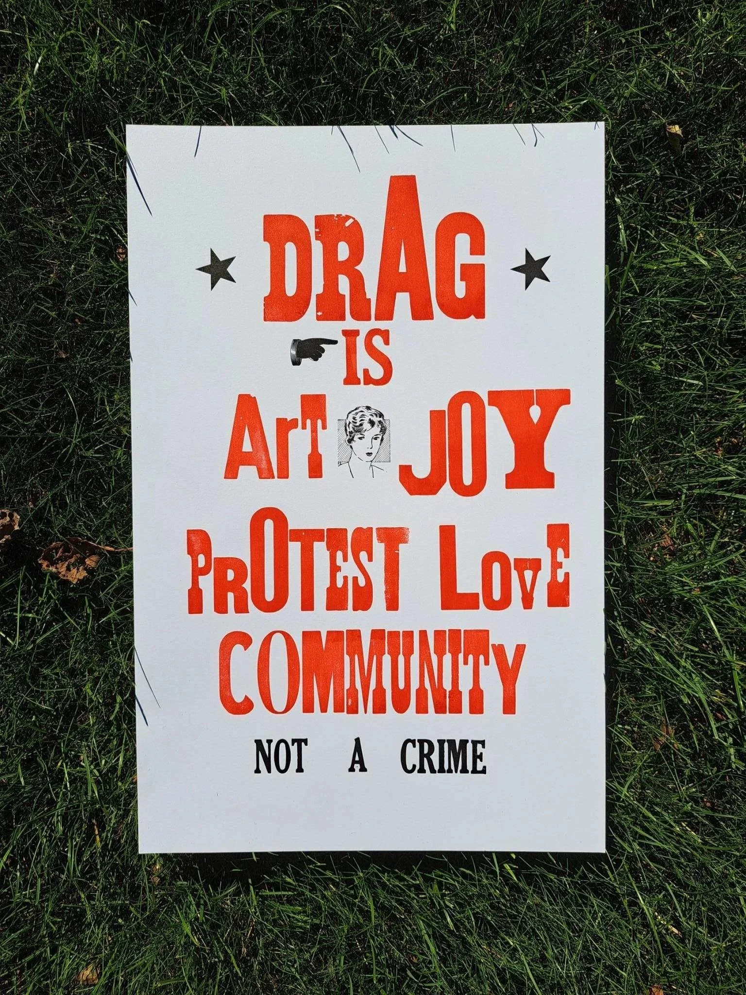 Letterpress poster displayed on grass reading “Drag is art, joy, protest, love, community, not a crime.”