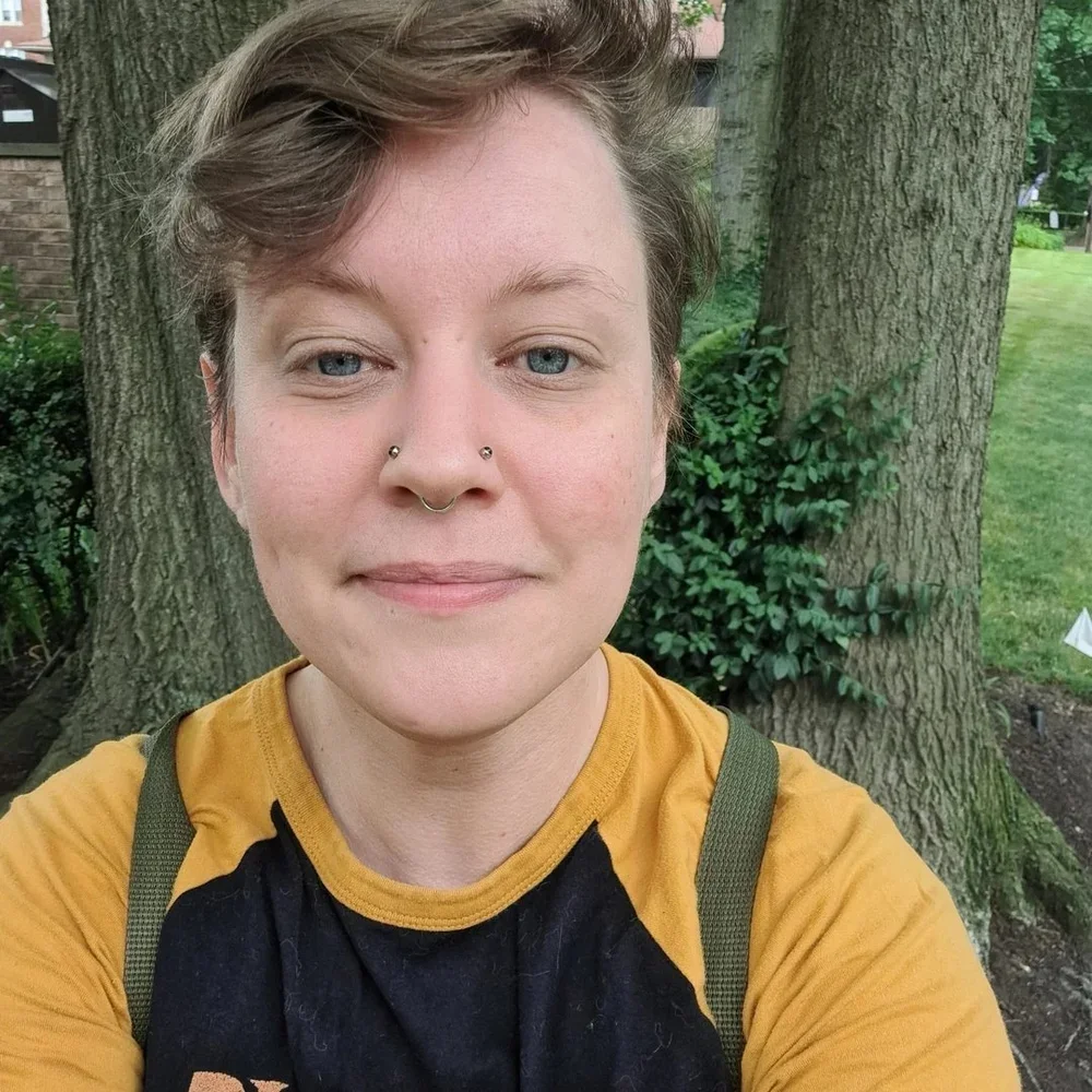 Portrait of Andy Passchier outdoors, smiling at the camera in a casual yellow-and-black top, standing in front of greenery and a tree.