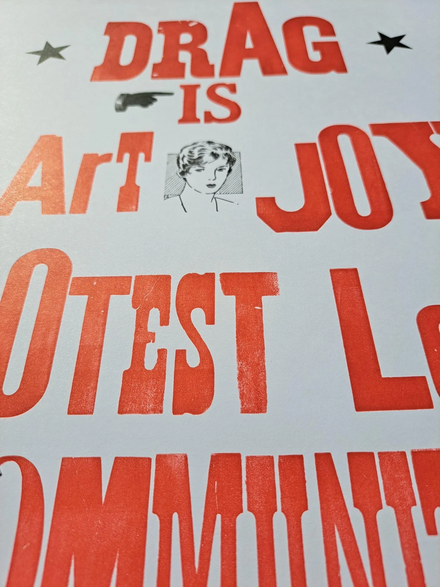 Close-up photograph of a red-and-black letterpress poster with oversized text reading “Drag is art, joy, protest, love, community.”