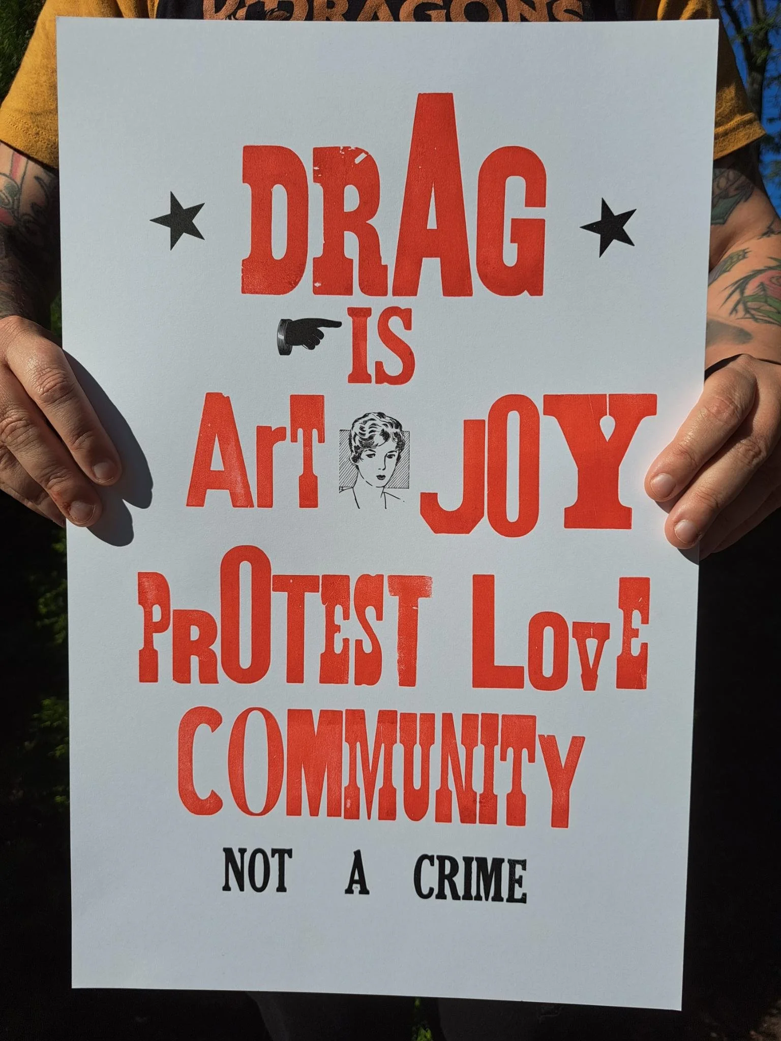 Person holding a red-and-black letterpress poster reading “Drag is art, joy, protest, love, community, not a crime.”