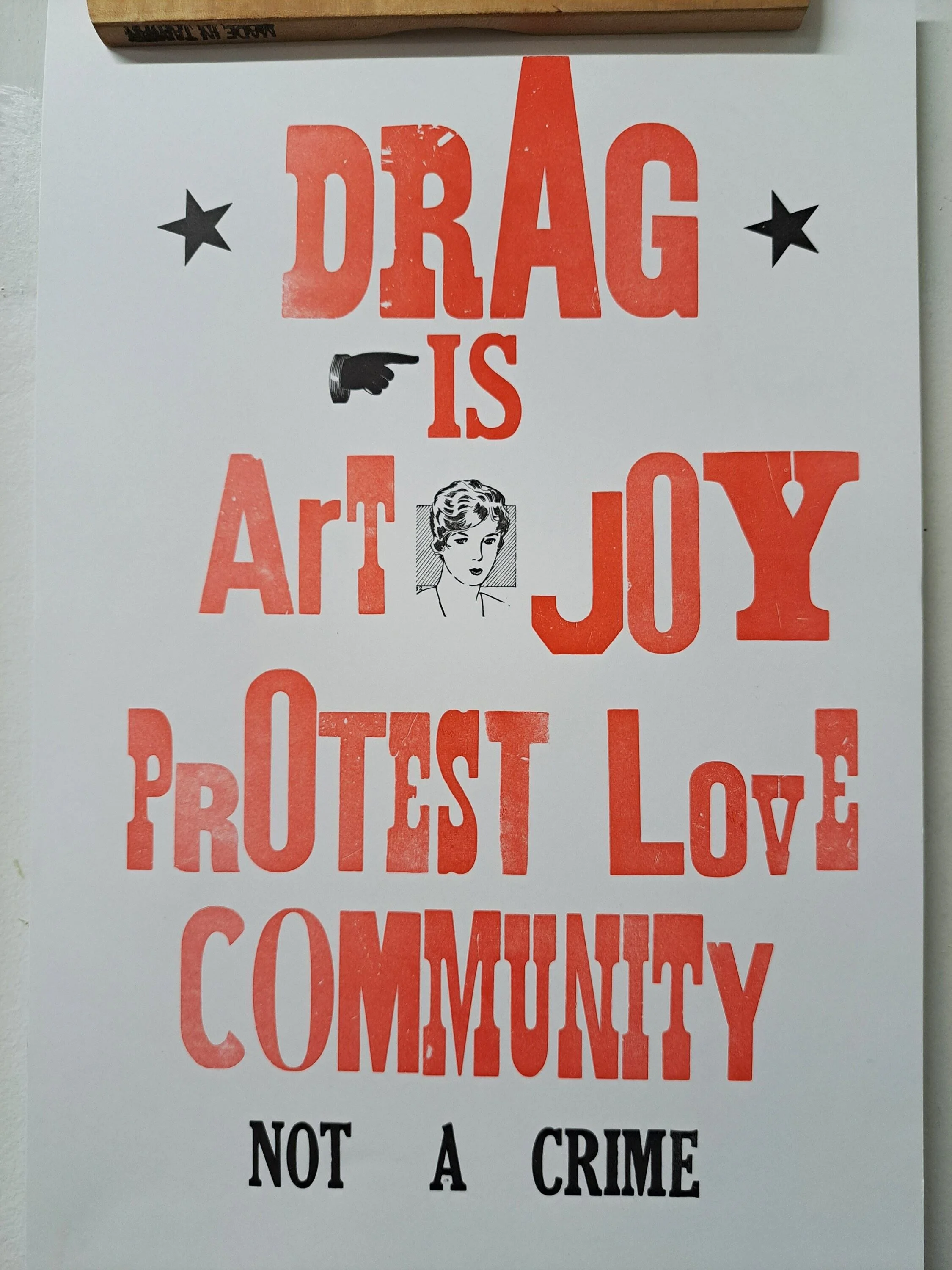 Close-up photograph of a red-and-black letterpress poster reading “Drag is art, joy, protest, love, community, not a crime.”