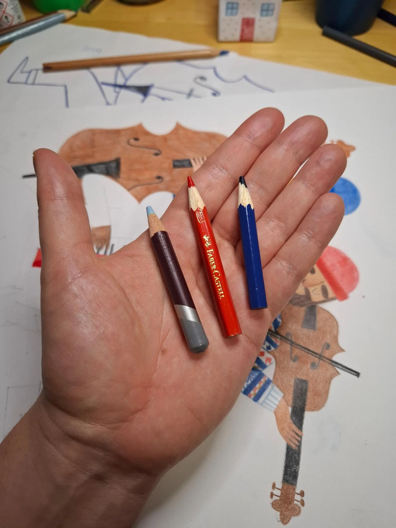 Process photo of a hand holding three colored pencils above a character illustration in progress on a drawing desk.