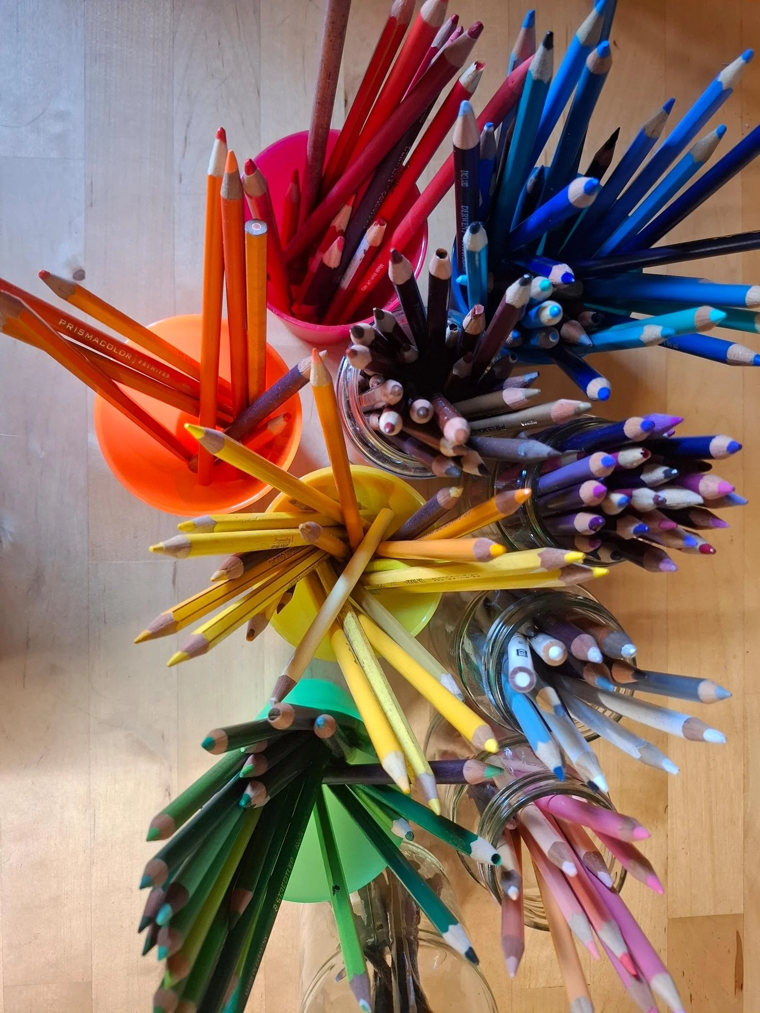 Studio photograph of colored pencils sorted by color in cups and glass jars on a wooden table.