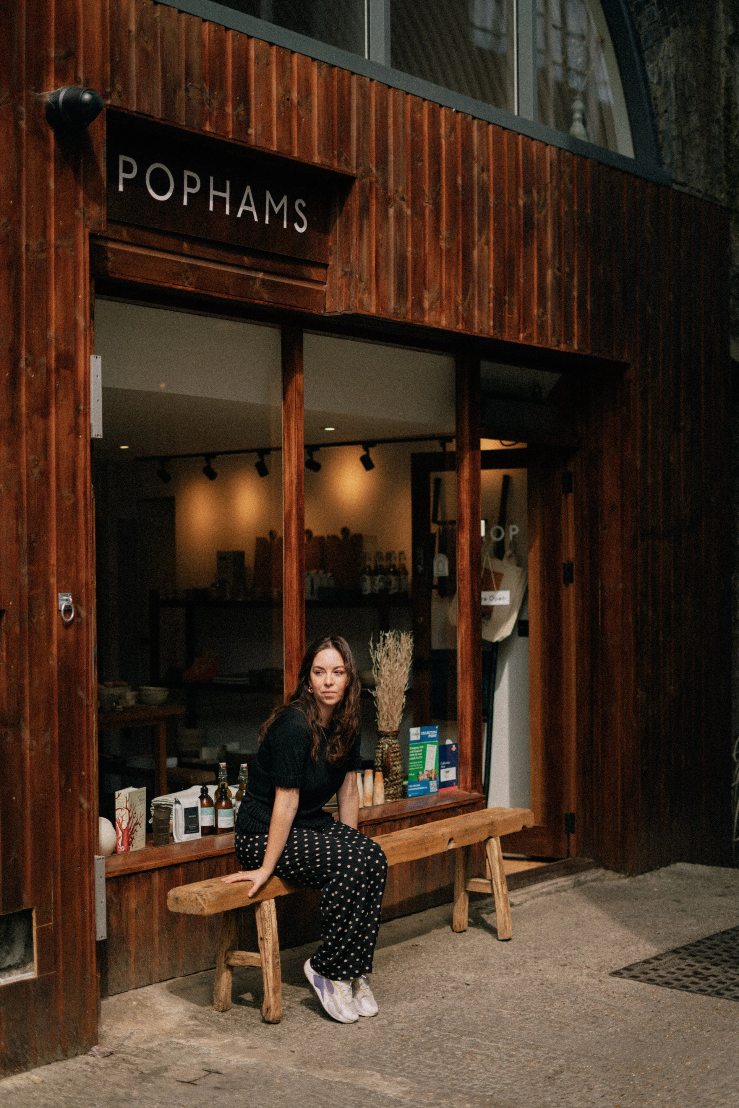 A woman sitting on a wooden bench outside a shop with a wood exterior and a window. She is wearing a black top, black and white polka dot pants, and white sneakers, with her left hand resting on the bench. The shop window displays bottles and promotional materials, and the shop sign reads 'POPHAMS'.