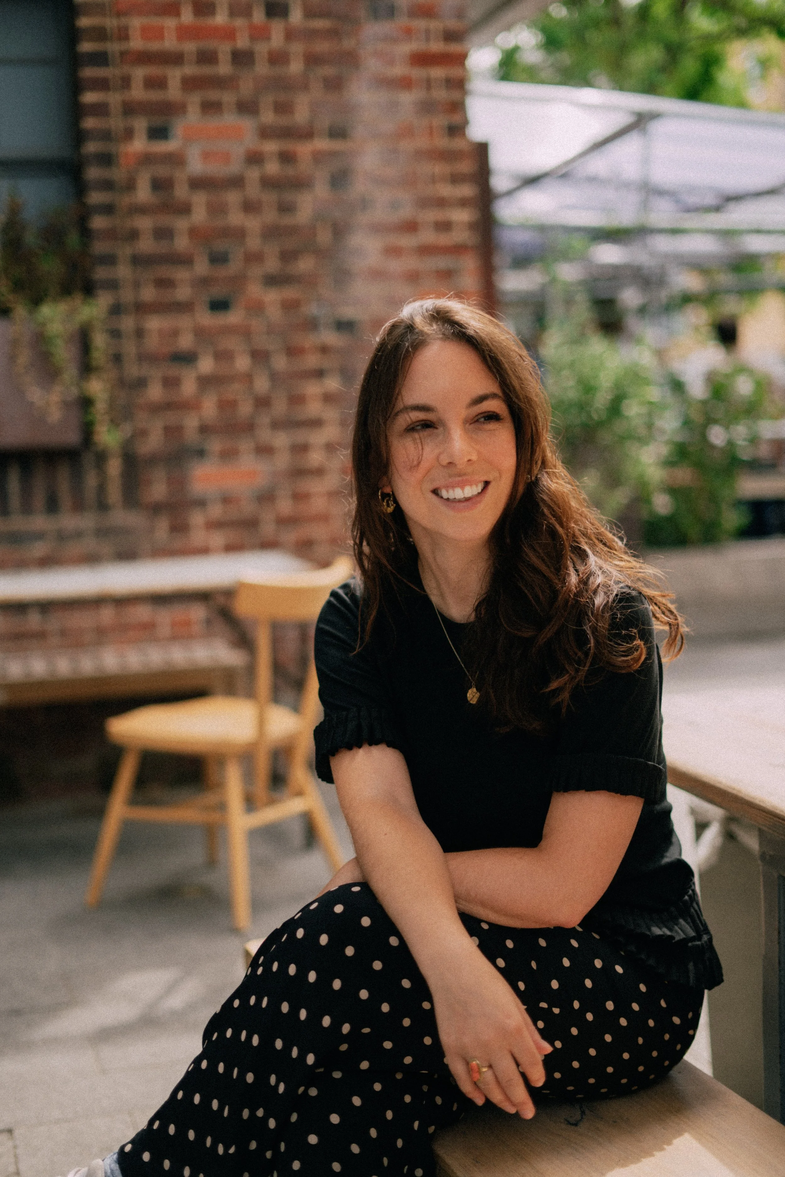 A woman with long brown hair, wearing a black top and polka dot pants, sitting outdoors at a wooden table, smiling and looking to her right, with a brick wall and green trees in the background.