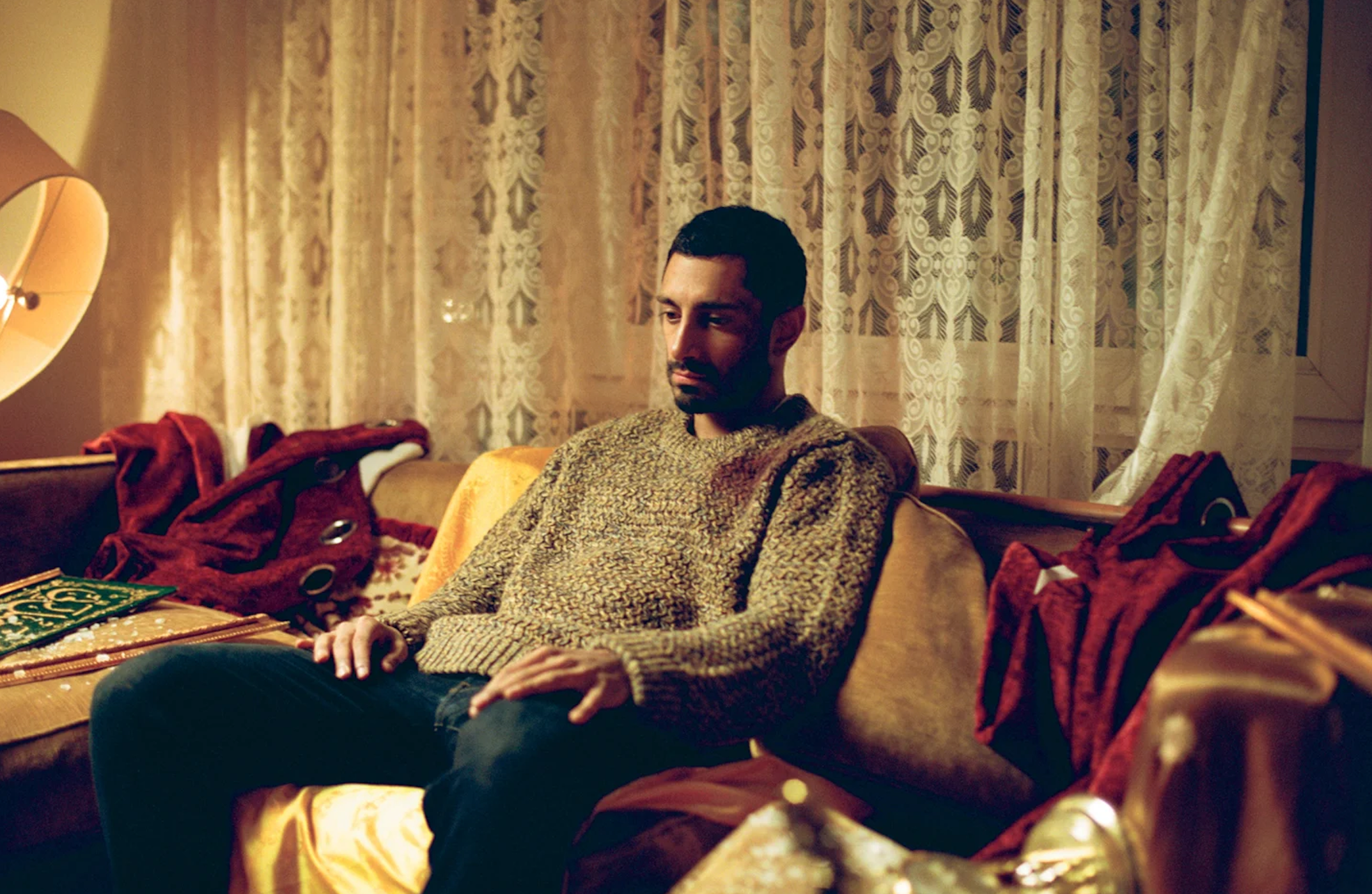 A man with short dark hair and a beard, wearing a patterned brown sweater, sitting on a yellow couch in a room with lace curtains, looking down with a neutral expression.