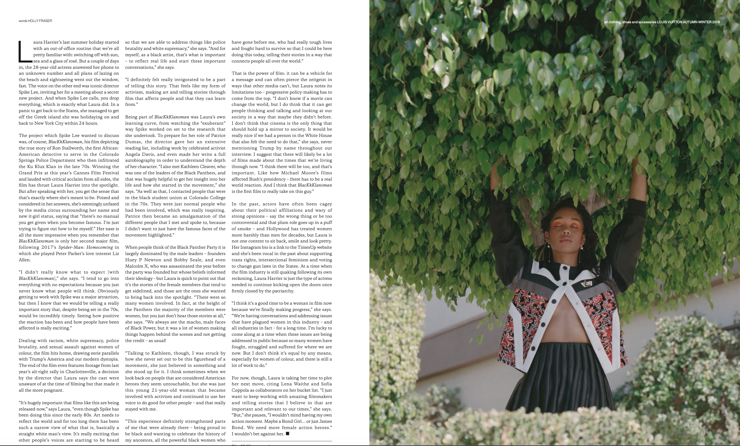 A woman with medium skin tone and black wavy hair is standing outdoors beneath a large bush with green leaves. She is wearing a black and white striped top, a patterned jacket, and a pleated black skirt. She has her eyes closed and one arm raised above her head.