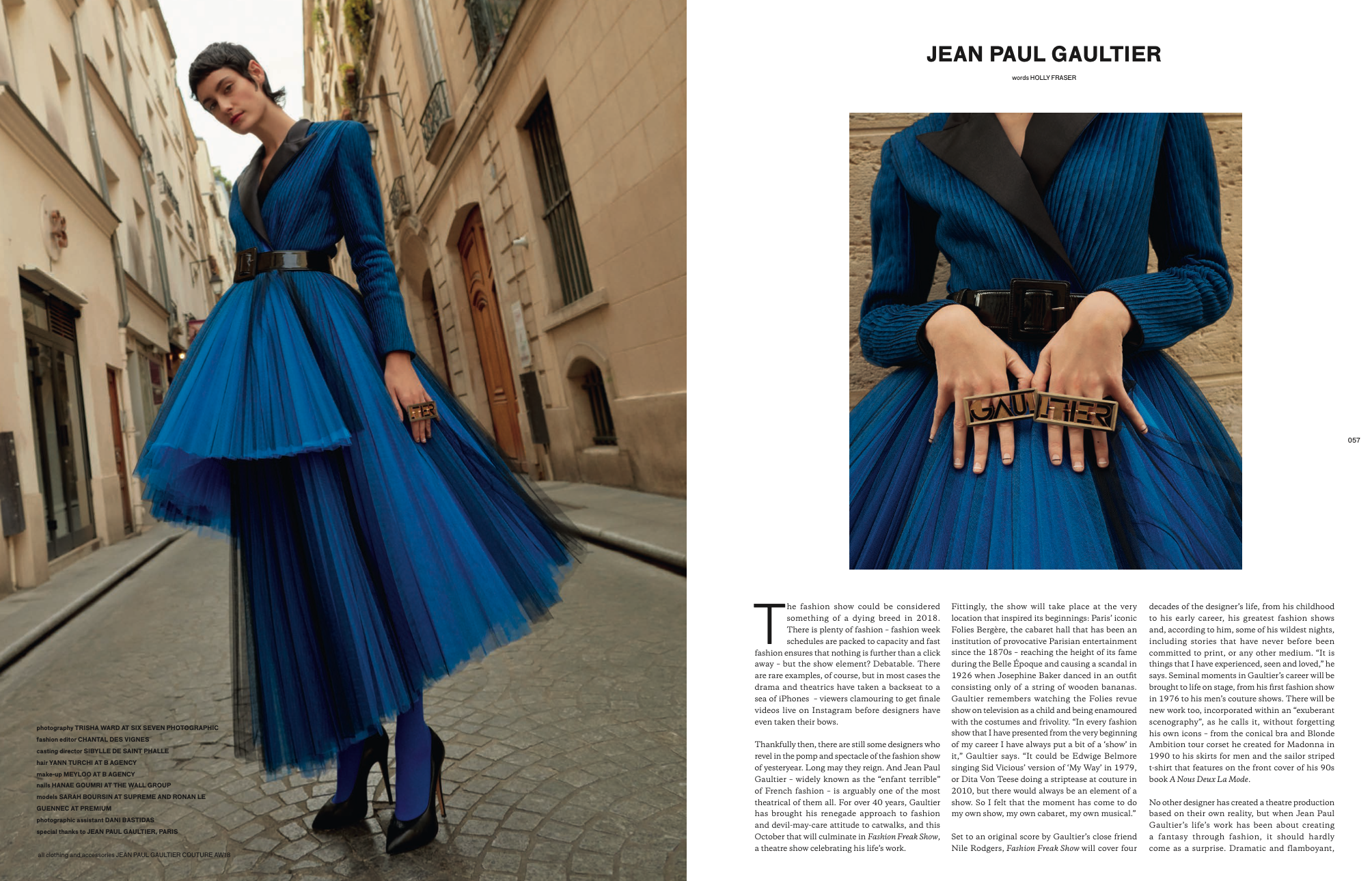 Fashion magazine spread featuring a woman in a blue pleated dress with black accents, standing on a cobblestone street in an urban setting, with a close-up of her hands holding a bold ring that spells 'GAULTIER.'