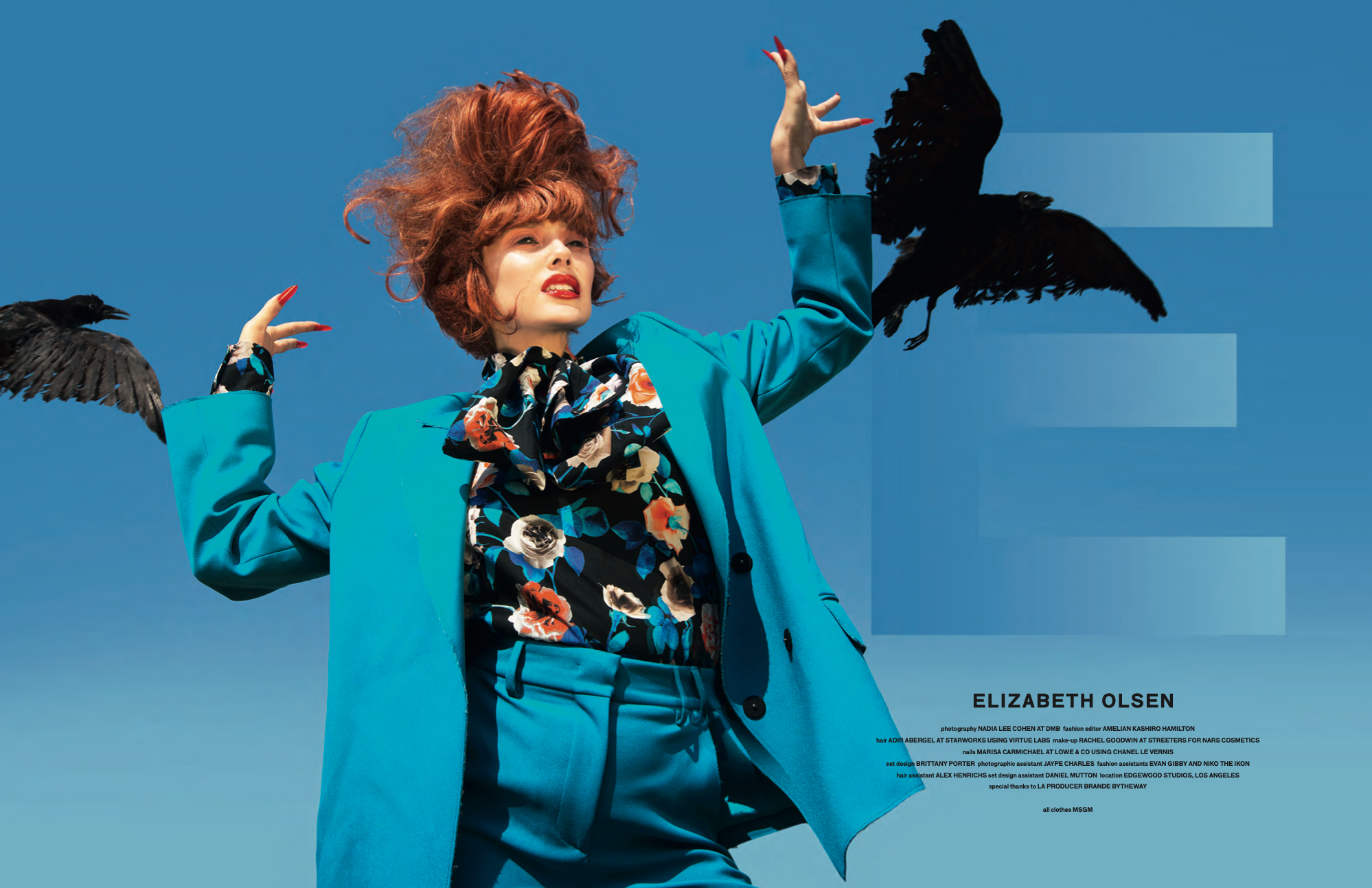 A woman with vibrant red hair styled in a wild, voluminous manner, wearing a bright blue blazer and a floral patterned shirt, is raising her hands with black birds flying around her against a clear blue sky background.