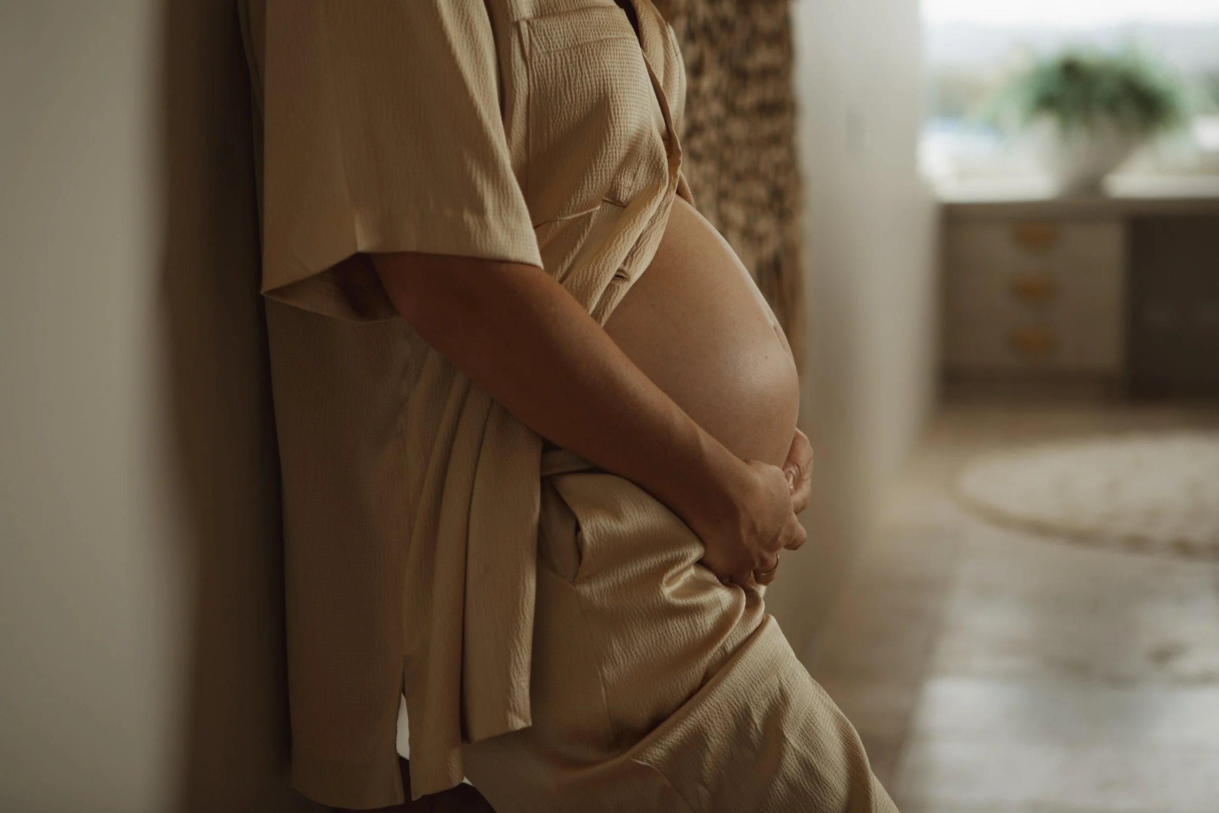 A pregnant person wearing a beige outfit is sitting on the floor, leaning against a wall in a sunlit room, with their hand resting on their baby bump.