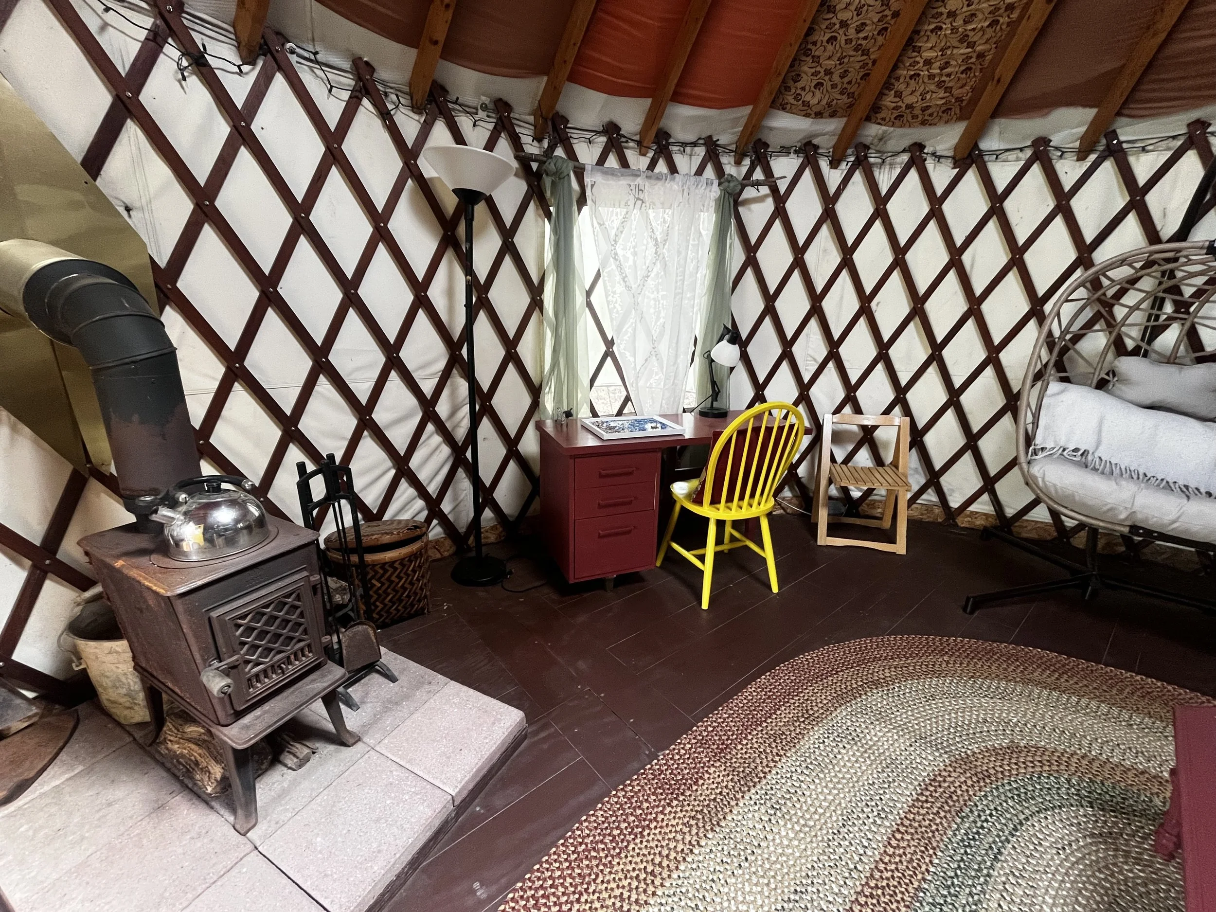 Interior of a yurt with a wood stove, a small desk, chairs, and a cozy seating area, with a round braided rug on dark wood flooring, and a window with sheer curtains.