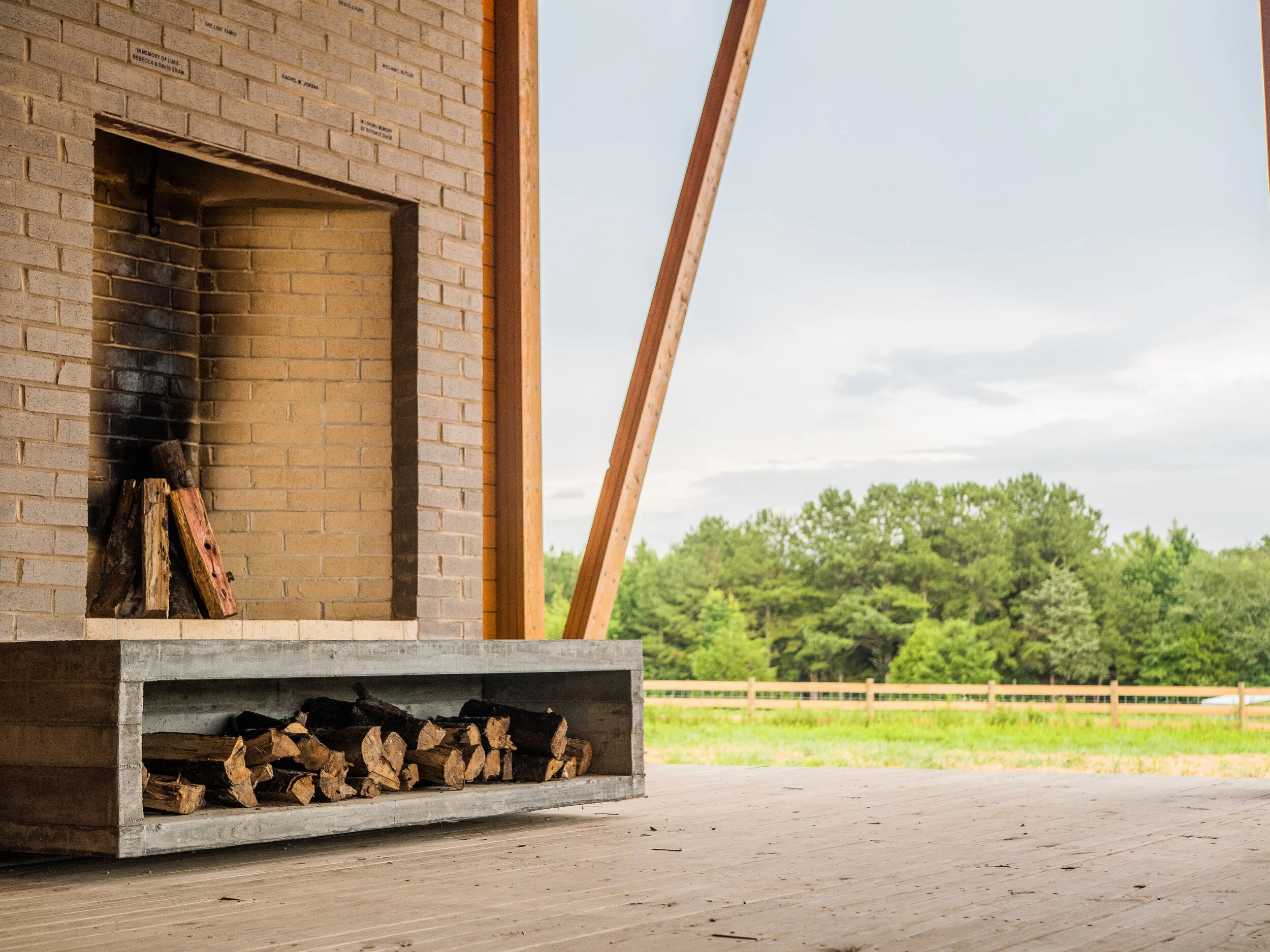 A brick fireplace with a wood storage area underneath and logs stacked inside. Construction appears to be ongoing with vertical wooden beams and a concrete base, set in an open outdoor area with green trees and a wooden fence in the background.