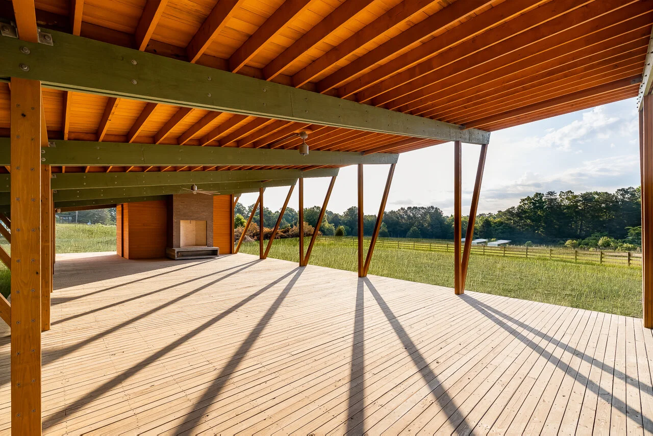 A spacious outdoor wooden deck under a modern architectural structure with angled supports, overlooking a grassy field and distant trees.