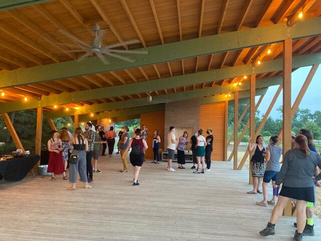People gathered at a social event inside a large, open pavilion with a high wooden ceiling, string lights, and wooden support beams, with some people dancing or talking.