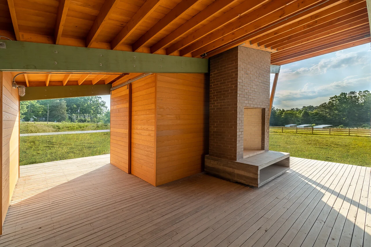 Wooden porch with a brick fireplace and open view of a grassy field and trees in the background.