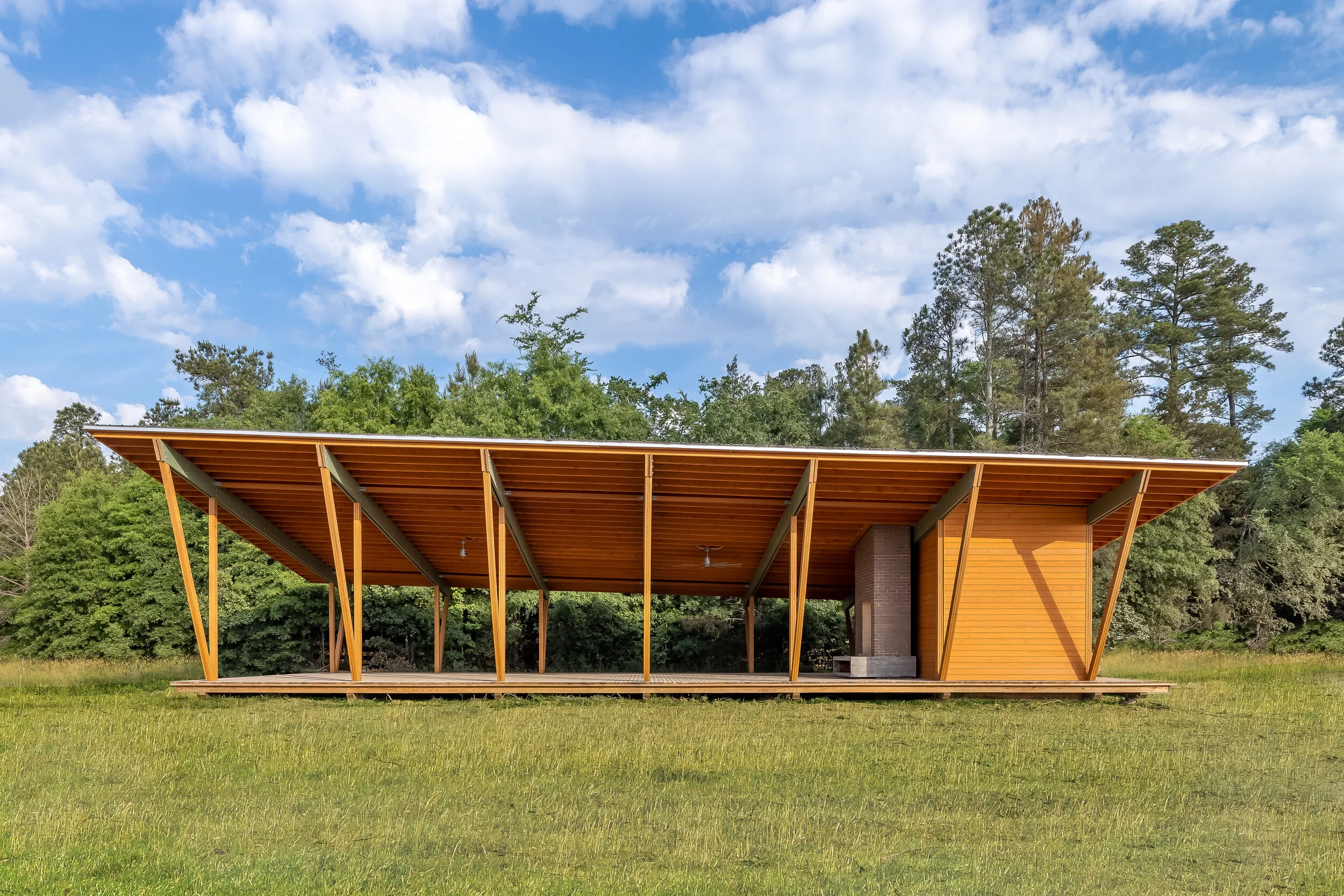 Modern open wooden pavilion with slanted roof supported by angled wooden beams, located in a grassy field with a background of green trees and a blue sky with scattered white clouds.
