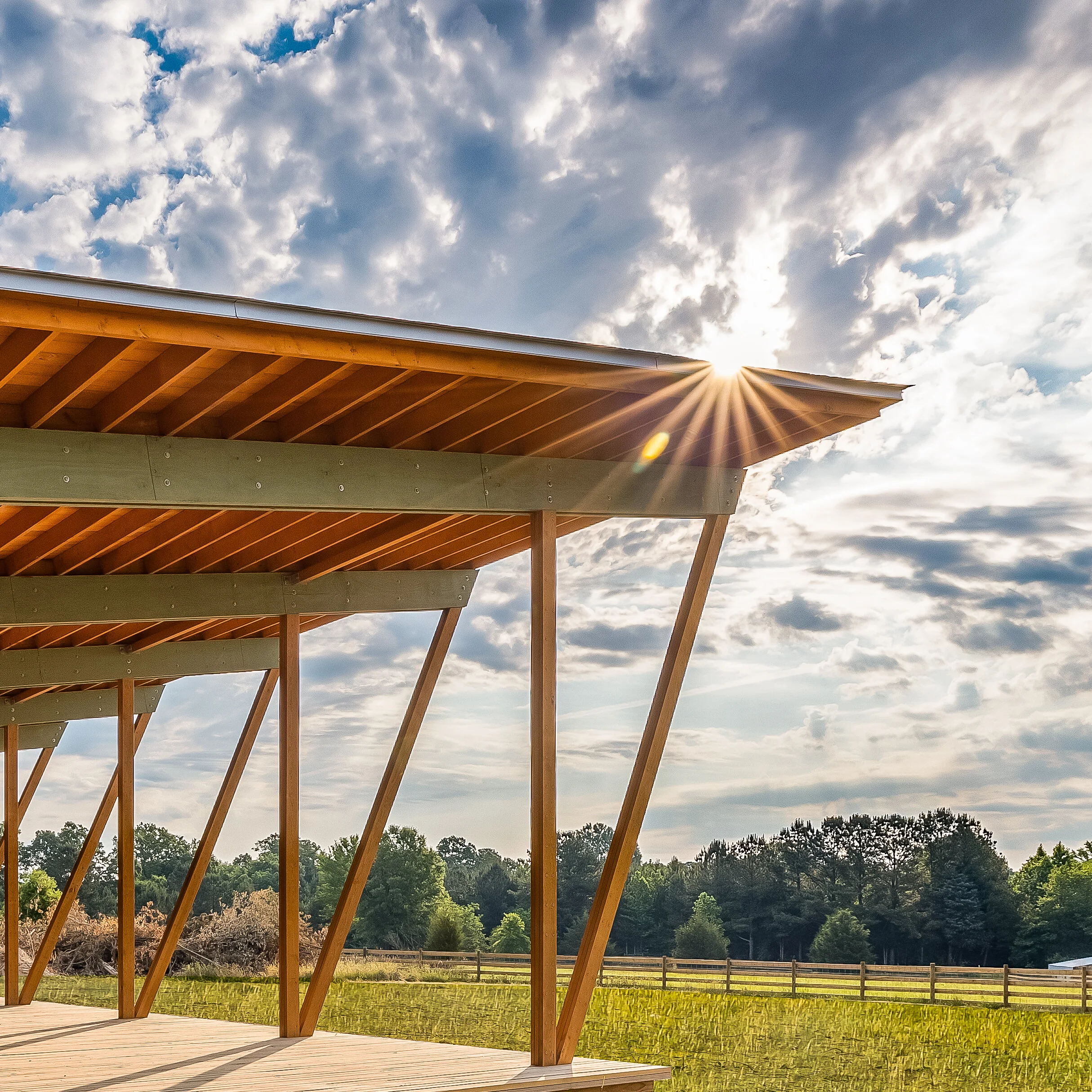 Wooden structure with a slanted roof under a partly cloudy sky with the sun shining behind the roof, overlooking a field and trees in the distance.