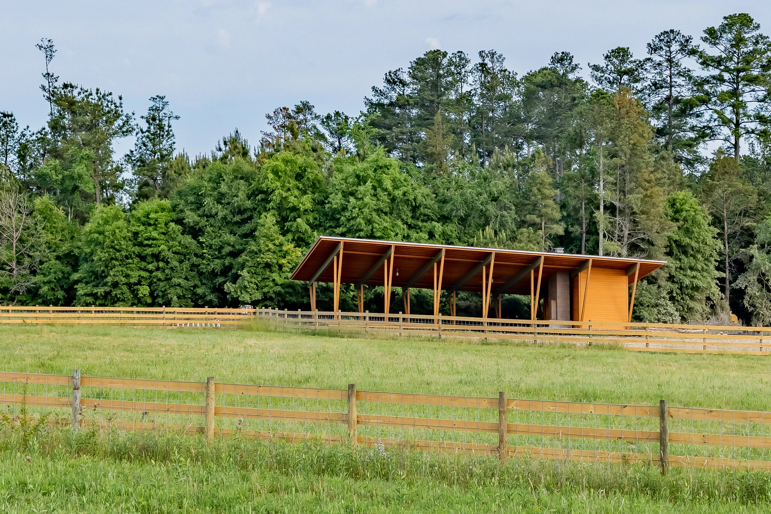 A modern wooden shelter with sloped roof stands in a green field with a wooden fence in the foreground. Tall trees and a partly cloudy sky are visible in the background.