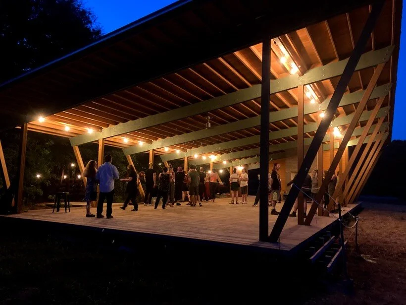 People gathered at an outdoor, open-sided wooden pavilion with warm lighting at dusk.