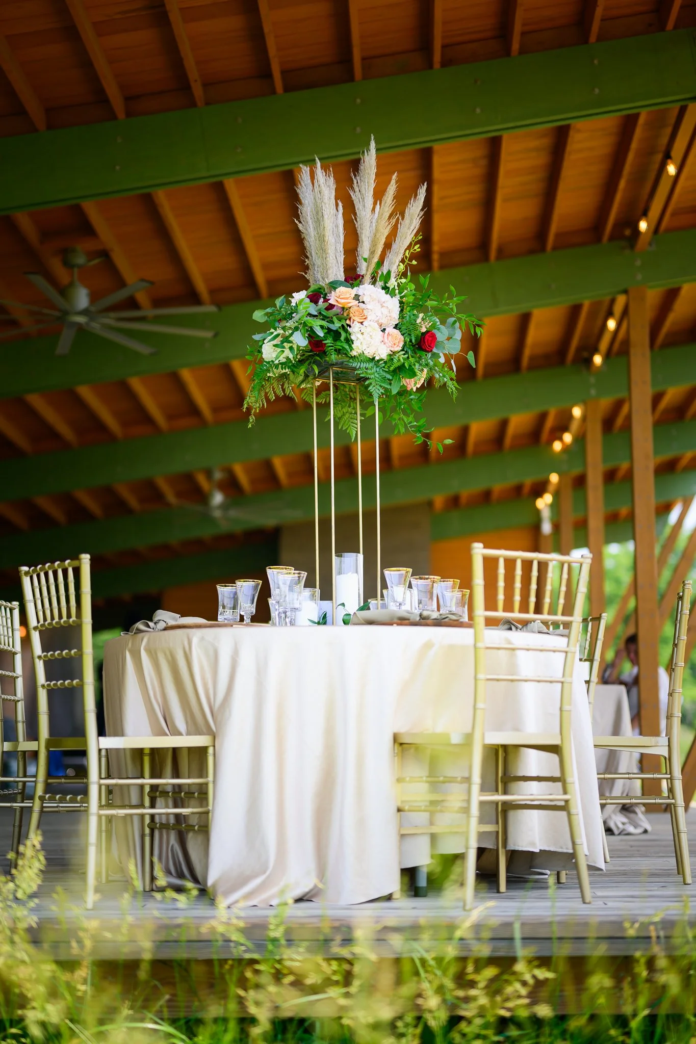 Table with a cream-colored tablecloth and gold chairs, decorated with a tall floral centerpiece of pampas grass, pink and red roses, and greenery, set in a rustic venue with wooden beams and green accents.