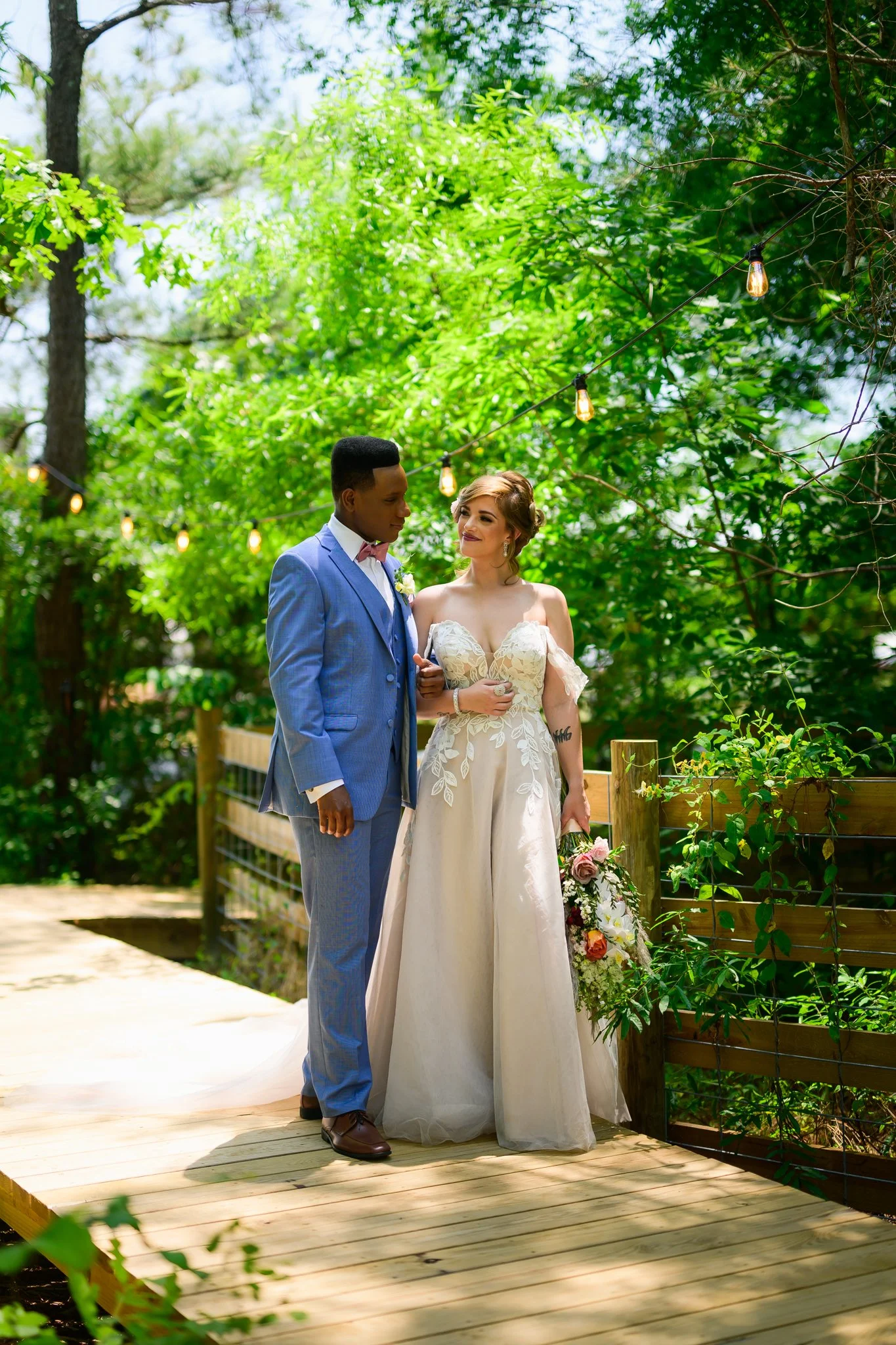 A bride and groom standing closely outdoors on a wooden walkway surrounded by green trees, with string lights above them, during their wedding.