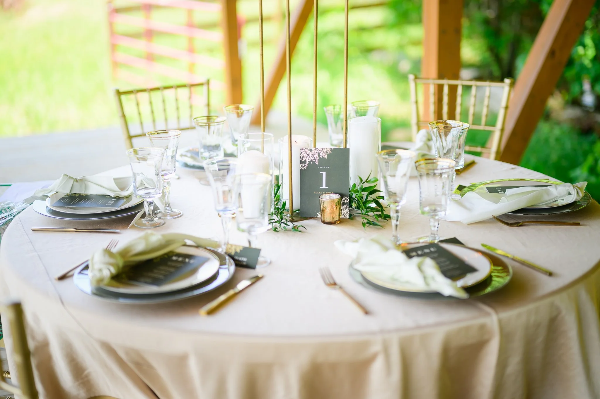 A round banquet table set for a formal event outdoors. The table is covered with a cream-colored tablecloth and has place settings with black placemats, white napkins, and gold utensils. There are multiple wine glasses at each setting. In the center 