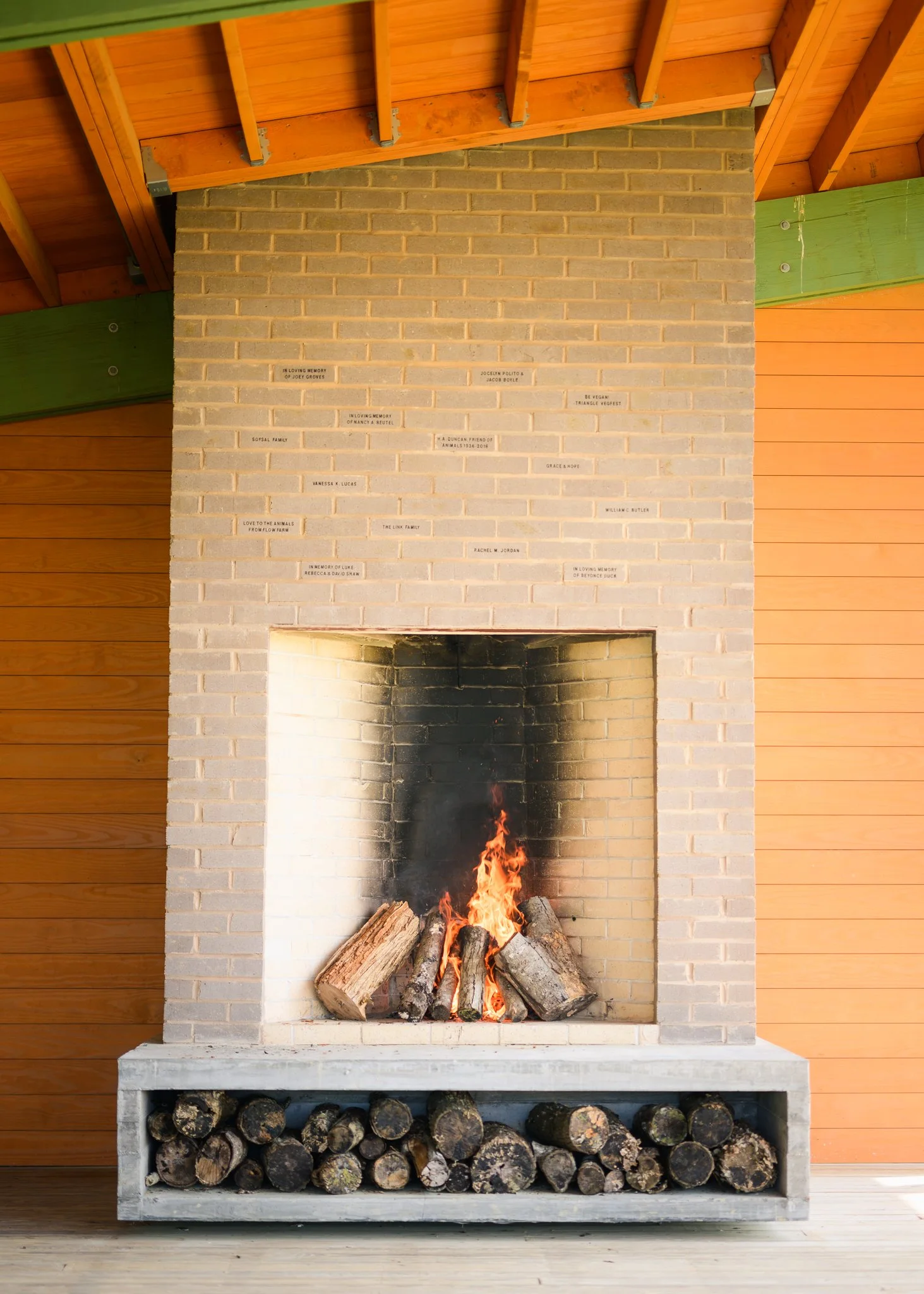 Fireplace with logs on the grate and flames, surrounded by a brick wall, with a wood storage area underneath and wooden paneling on the sides.