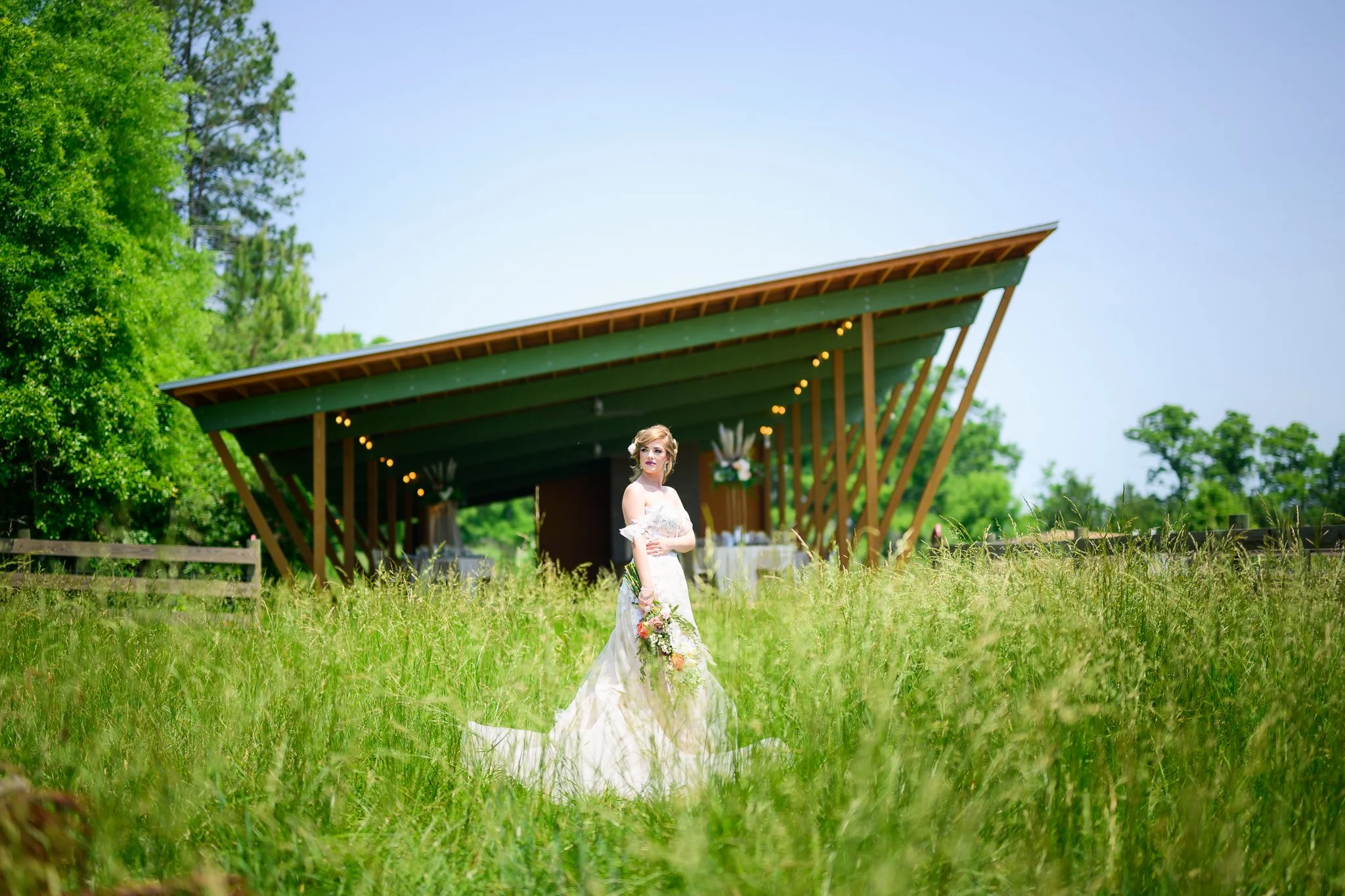 A woman in a white wedding dress holding a bouquet, standing in a grassy field during a wedding ceremony with a modern outdoor pavilion in the background.