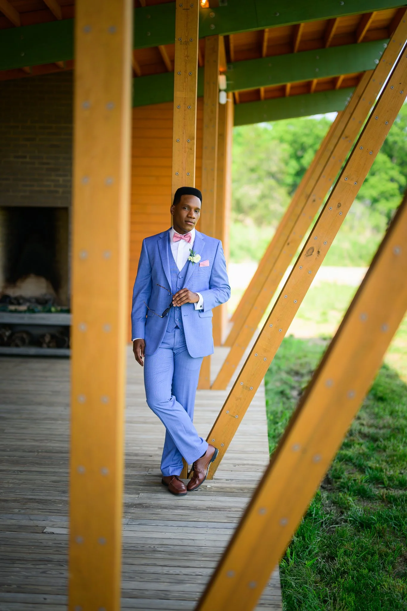 A man in a light blue suit, pink bow tie, and brown shoes standing outdoors on a wooden walkway, holding glasses, with wooden and green beams overhead and greenery in the background.