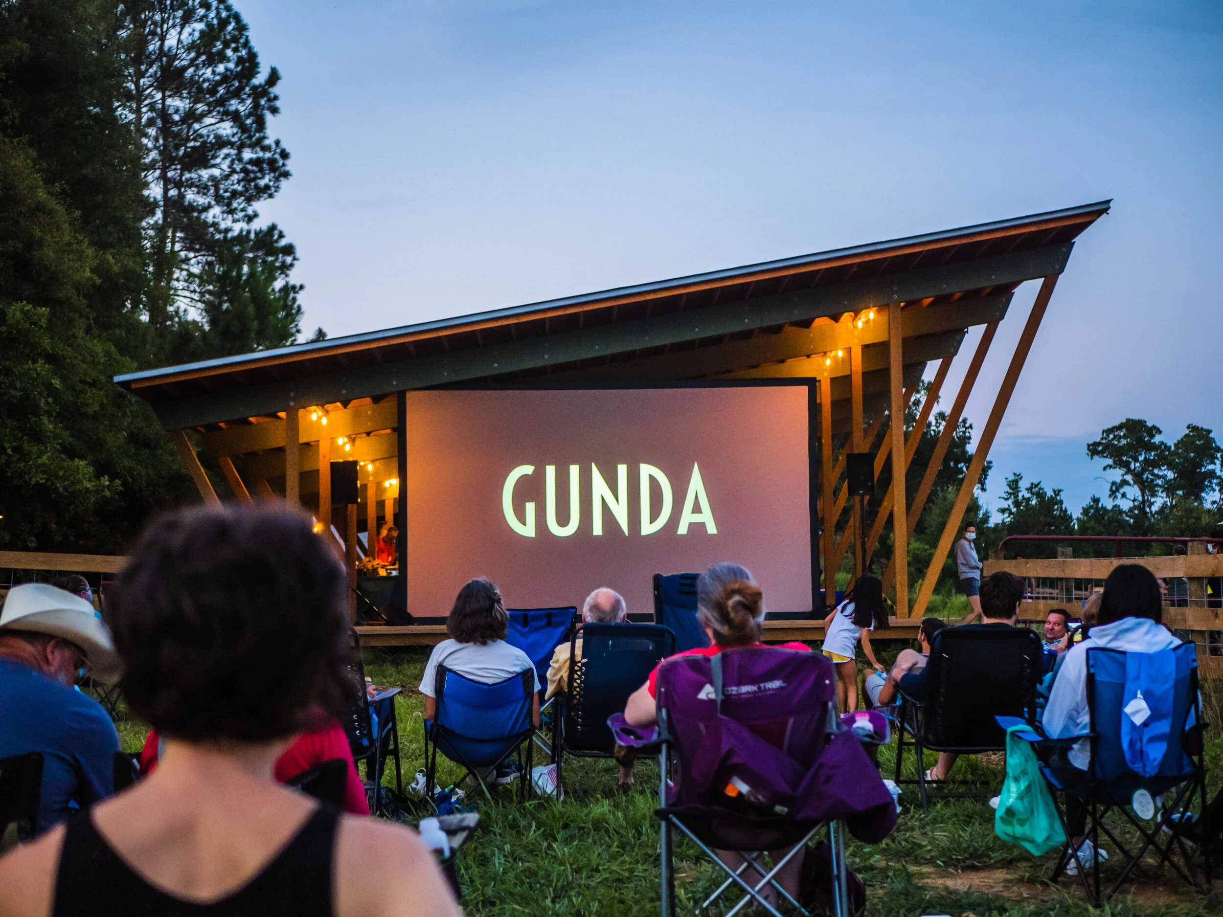People watching an outdoor movie screening at dusk, seated on chairs and surrounded by trees, with a large screen displaying the words 'CINDA GUNDA' and string lights hanging above the stage.