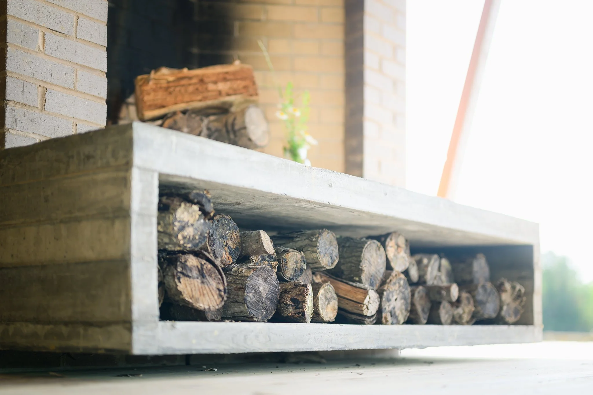 Stacked firewood logs stored under a concrete shelf with a brick wall background.