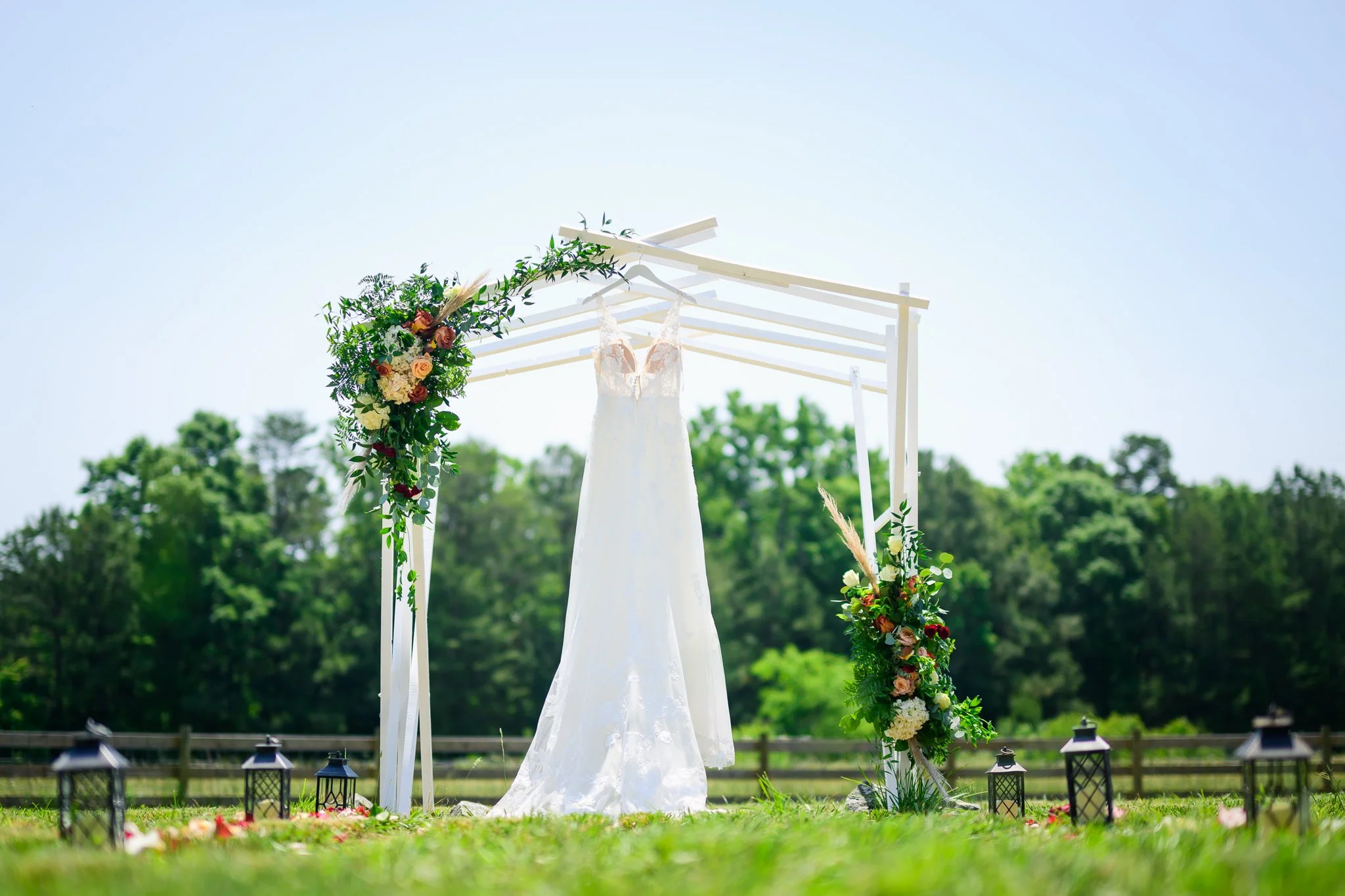 An outdoor wedding altar decorated with a floral arrangement and a wedding dress hanging in the center, along with small lanterns on the grass.