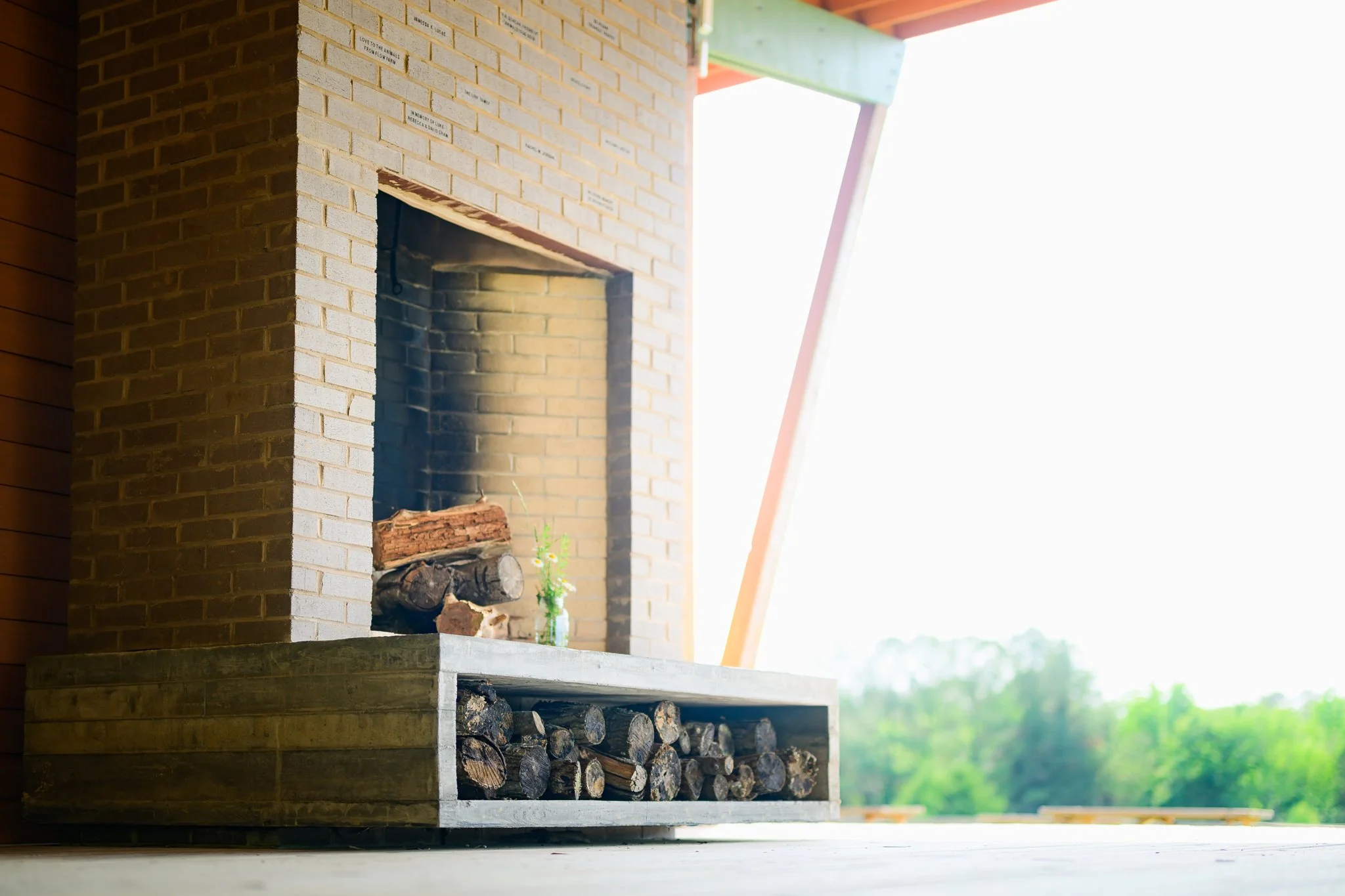 A brick outdoor fireplace with stacked firewood underneath, a small plant growing nearby, and part of a building with green and pink beams in the background.