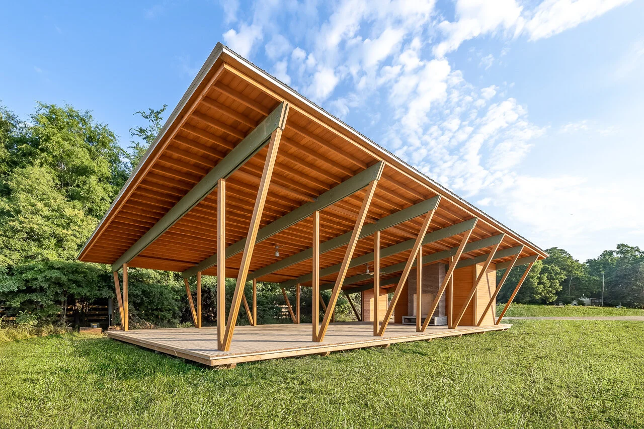 Modern wooden open-air pavilion with angled support beams and a raised deck, situated on a grassy area with trees in the background and a partly cloudy sky.